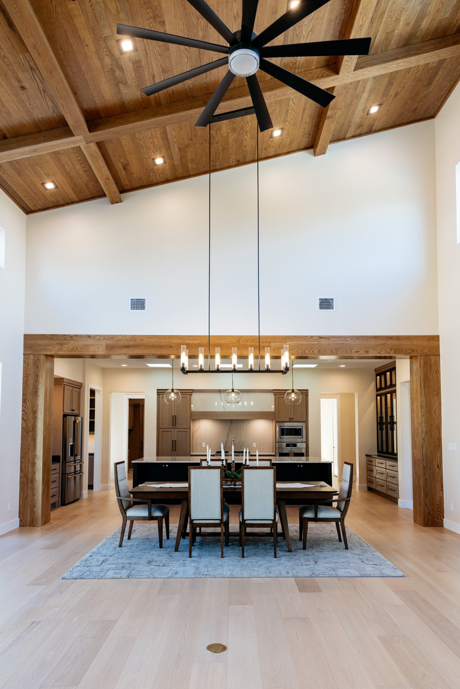 Open-concept kitchen and dining area with wooden ceiling beams, modern black chandelier above a large wooden dining table with eight chairs, and a kitchen with gray cabinets, stainless steel appliances, and a white marble island at the back. Light wo
