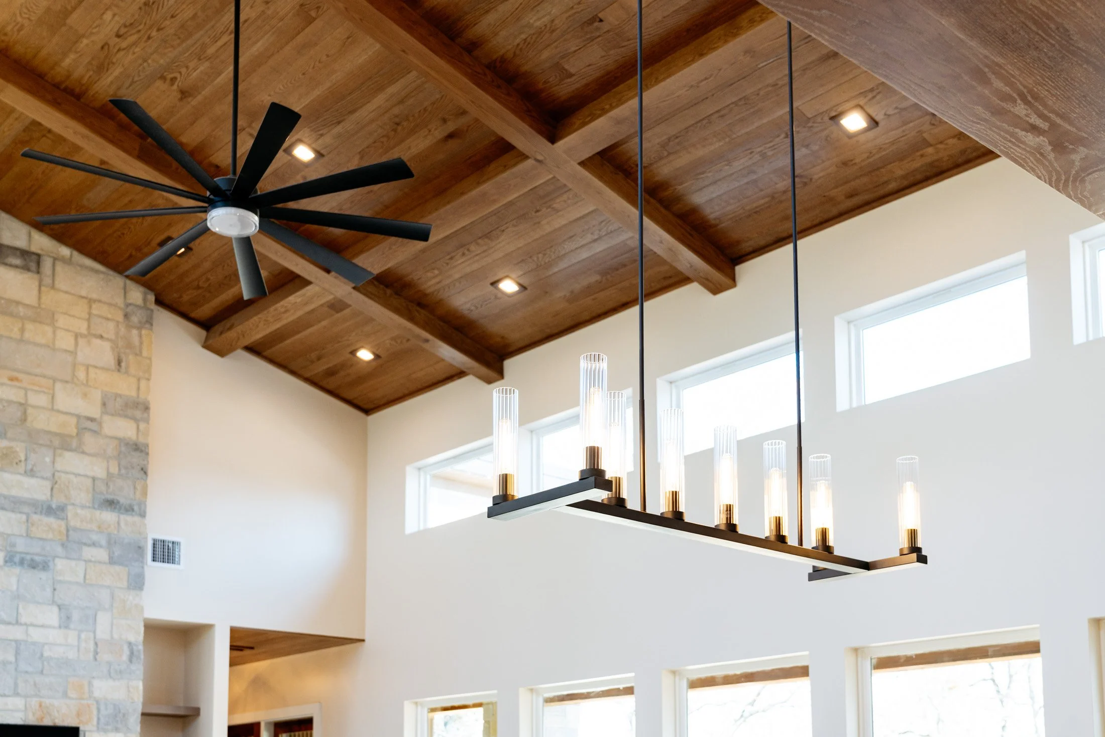 A modern interior ceiling with wood paneling, recessed lighting, a black ceiling fan, and a rectangular chandelier with multiple bulbs hanging above a large window.
