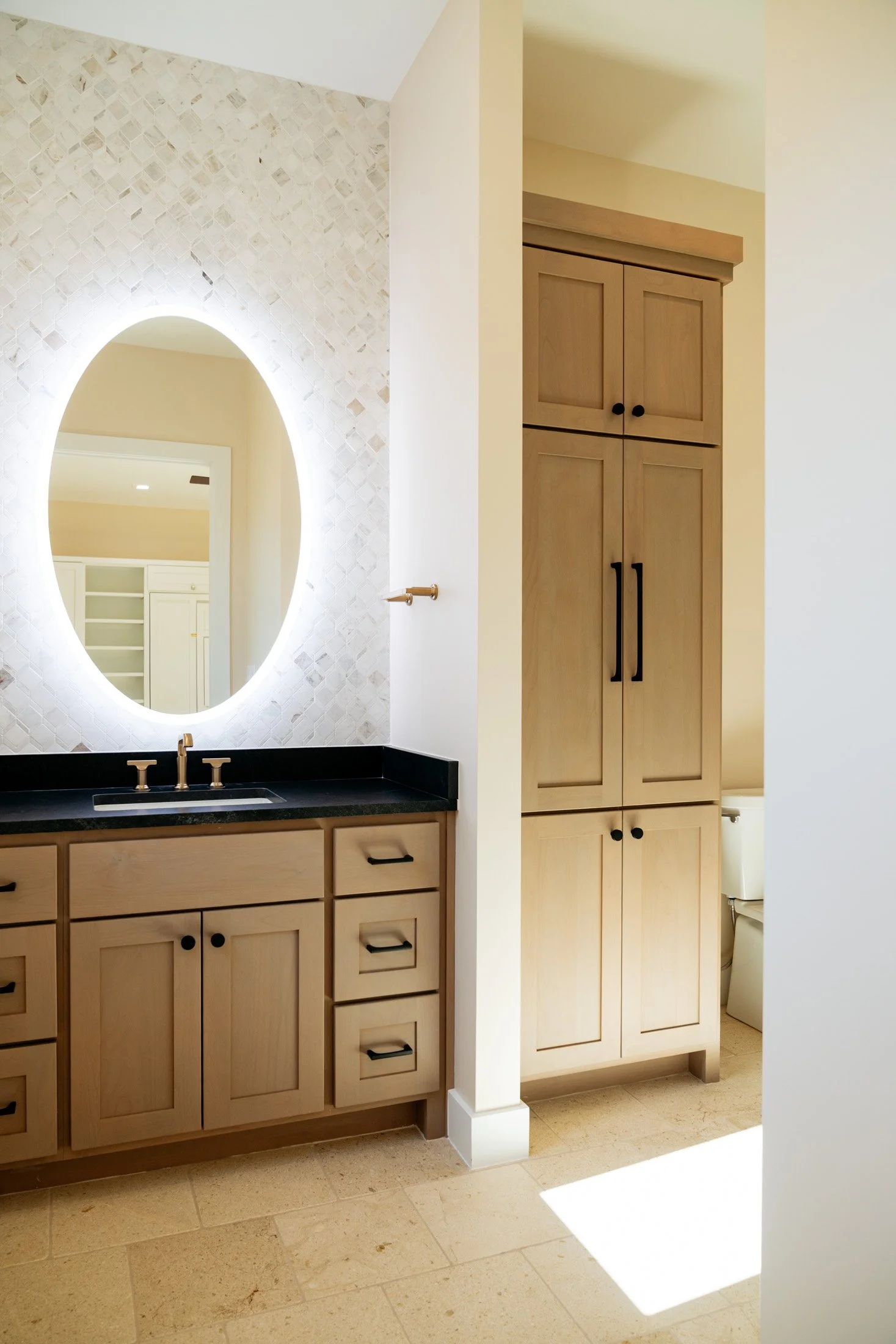 A bathroom vanity with a black countertop, a mirror with backlit lighting, and light wood cabinetry with black hardware. There is a bathroom cabinet on the right side.