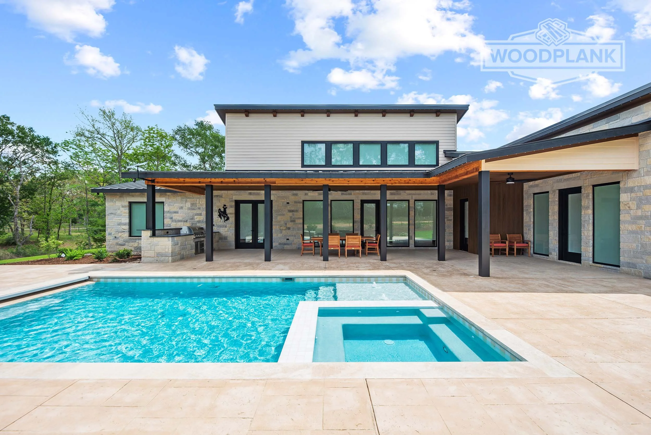 Modern house with stone and siding exterior, large glass doors, outdoor patio with wooden dining table and chairs, swimming pool in the foreground, surrounded by green trees and a blue sky with clouds.