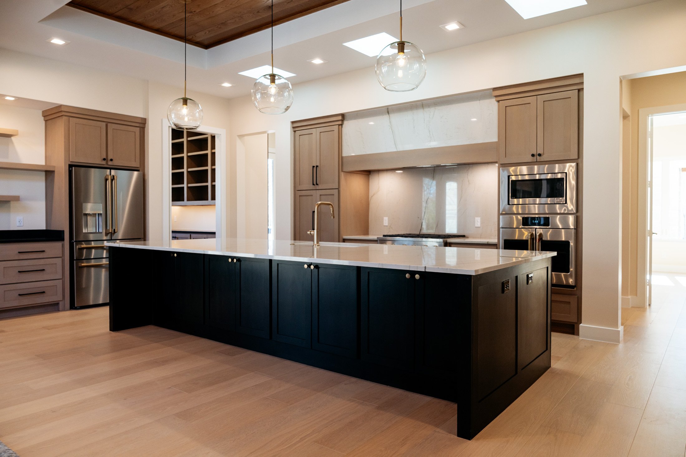 Modern kitchen with a large black island, light wood cabinetry, stainless steel appliances, and pendant lights hanging from a wooden ceiling.
