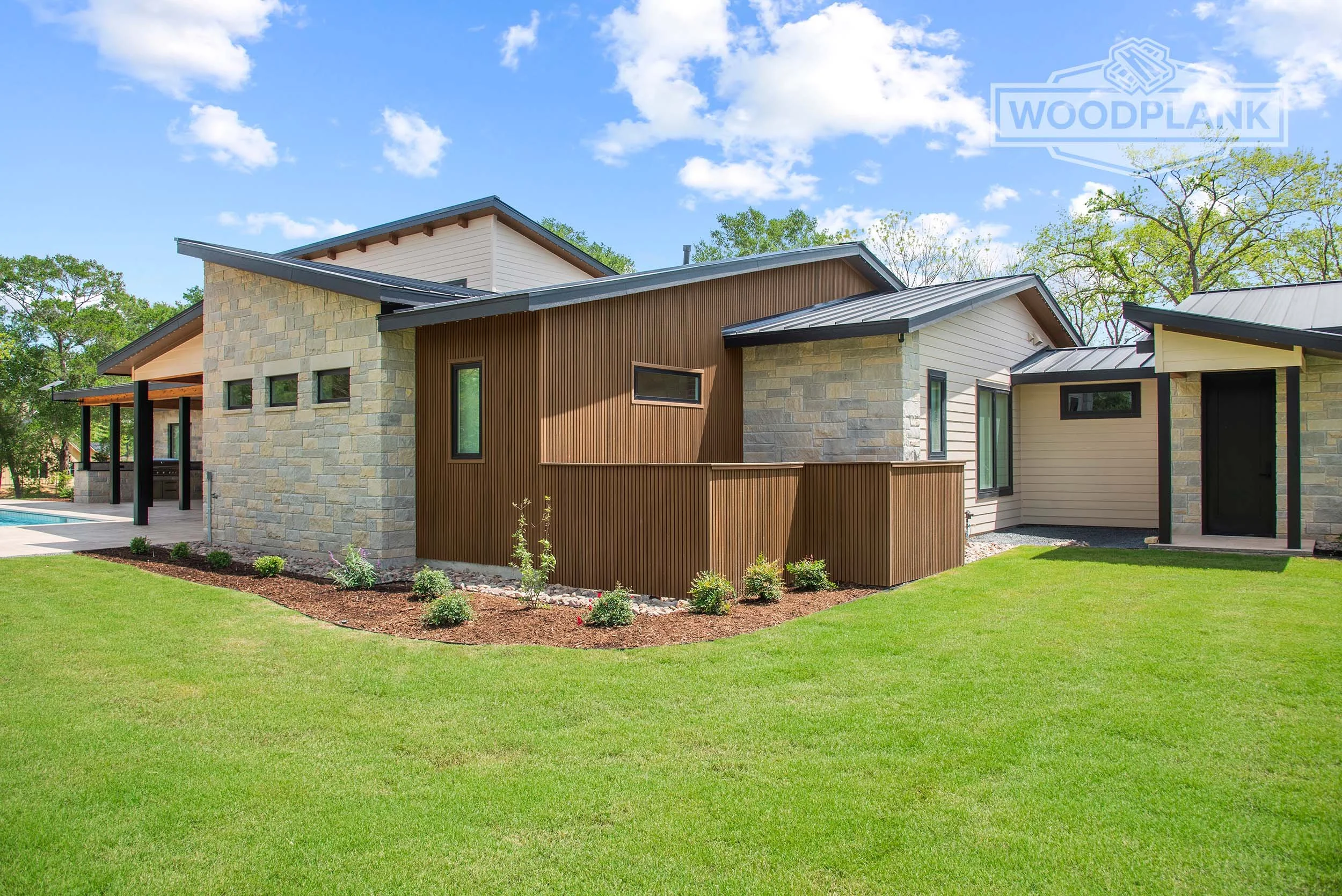 Modern house with stone and wooden exterior walls, large windows, and a metal roof, surrounded by a green lawn and trees under a partly cloudy sky.