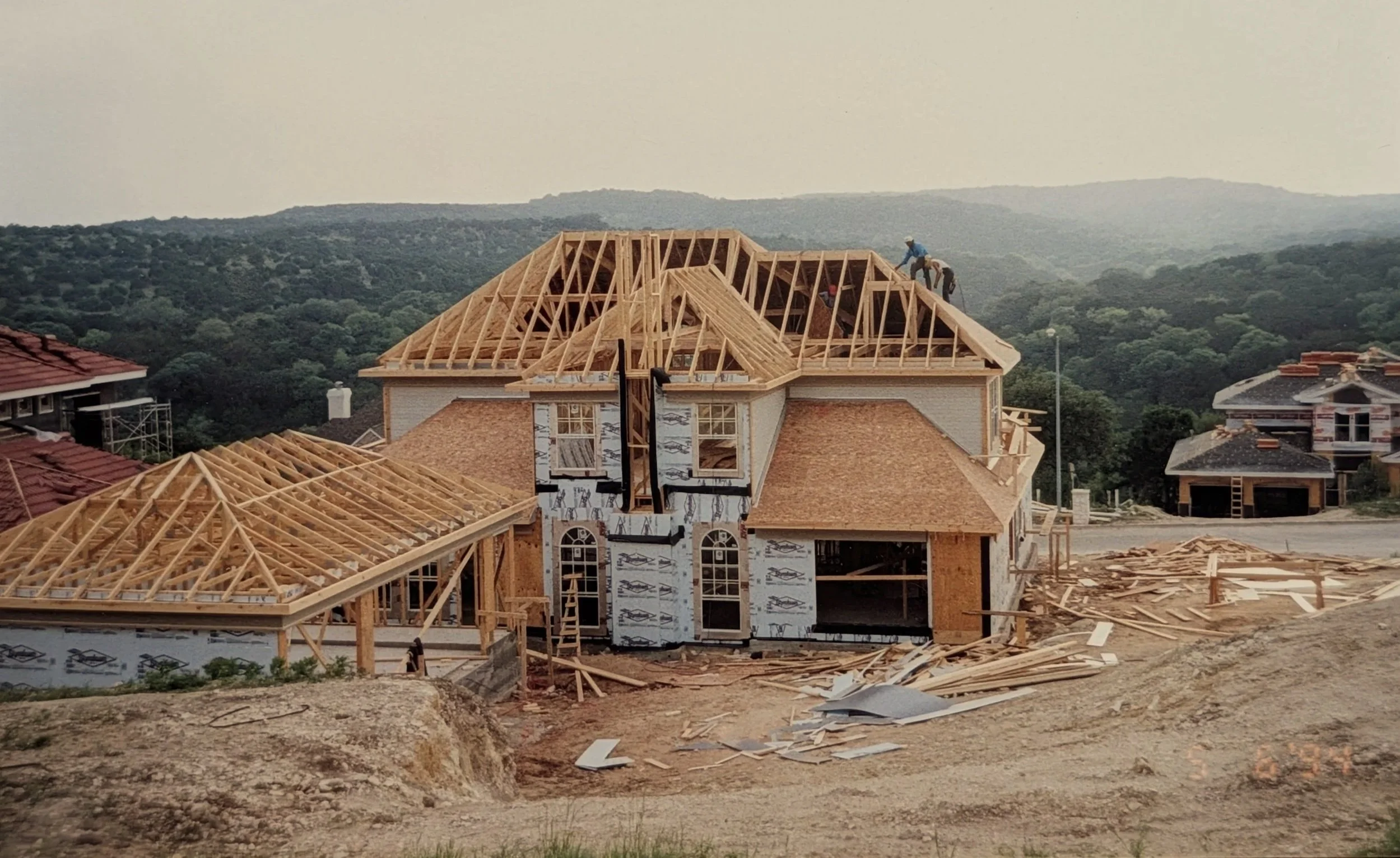A house under construction with a wooden roof frame, scaffolding surrounding it, and workers on the roof, with a scenic landscape of hills and trees in the background.