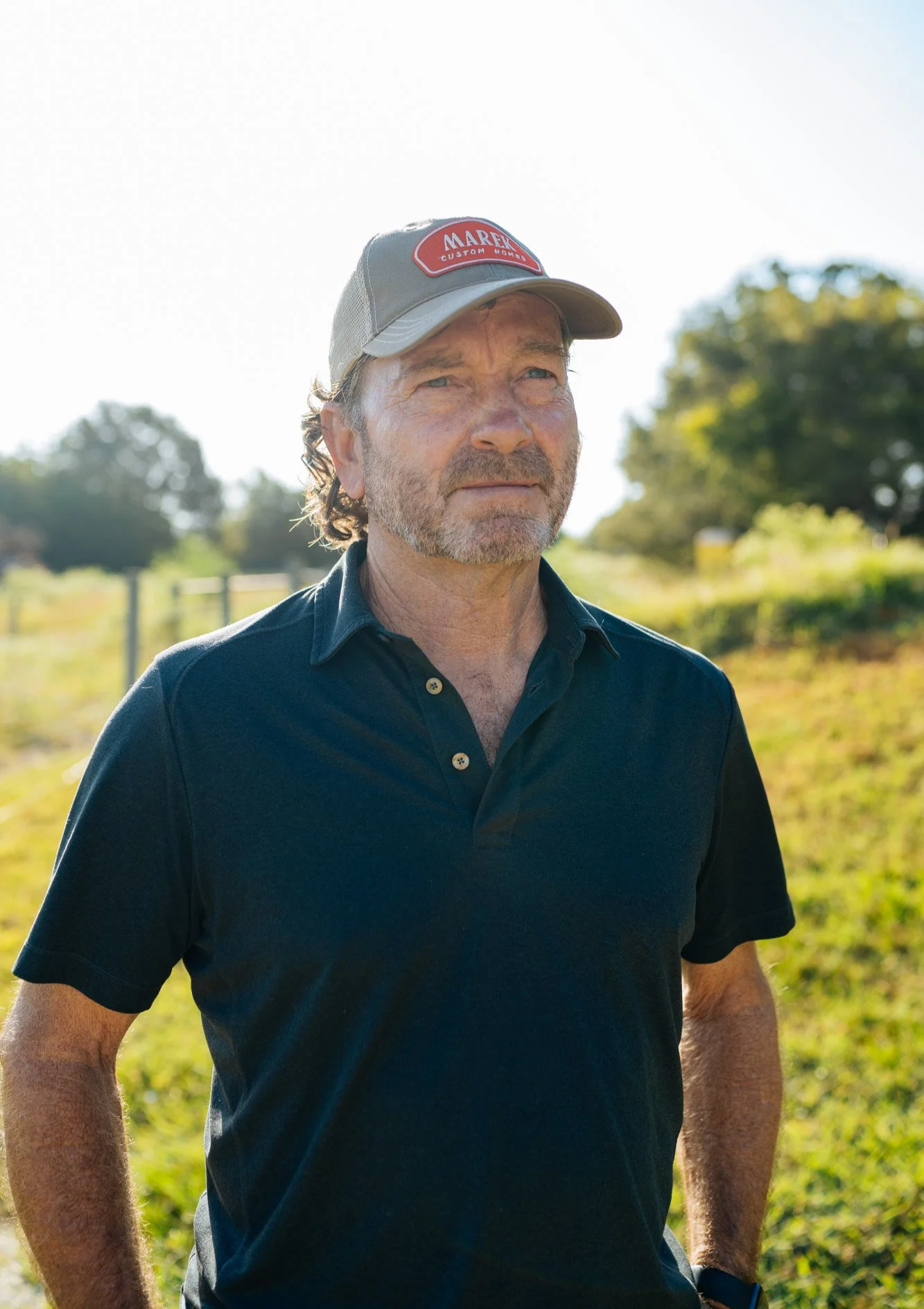 A middle-aged man with grayish hair and beard wearing a gray baseball cap with red and white patch that says "MAREK CUSTOM HOMES" and a black polo shirt, standing outdoors in bright sunlight with a blurred background of green trees and grass.
