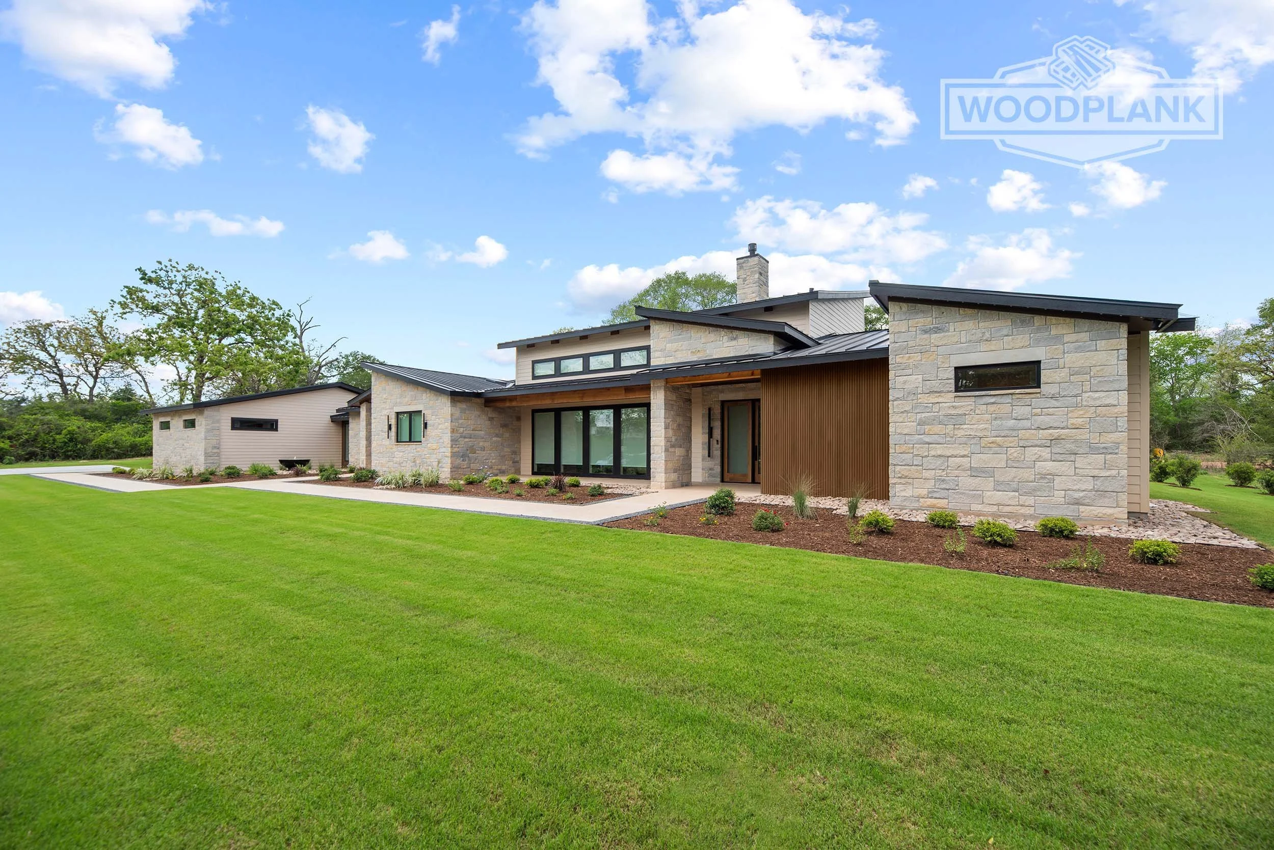 Modern house with stone and wood exterior, surrounded by a well-manicured lawn and trees, under a blue sky with clouds.