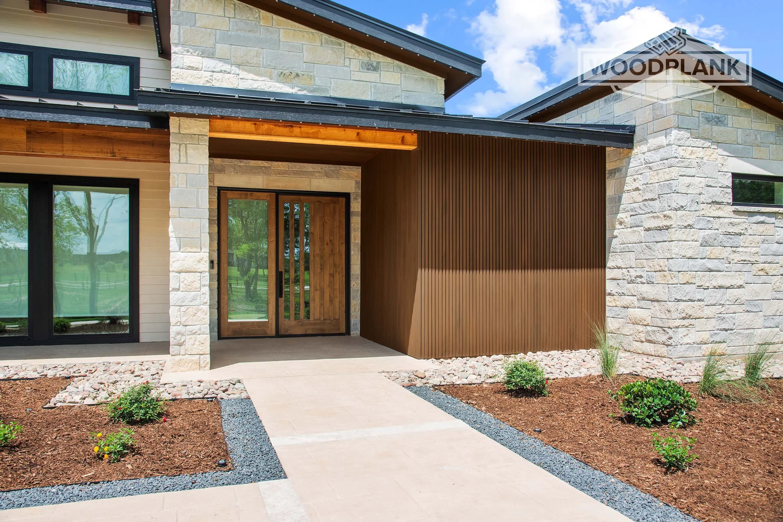 Modern house exterior with stone and wood siding, black window frames, and a landscaped front yard with small plants and mulch.