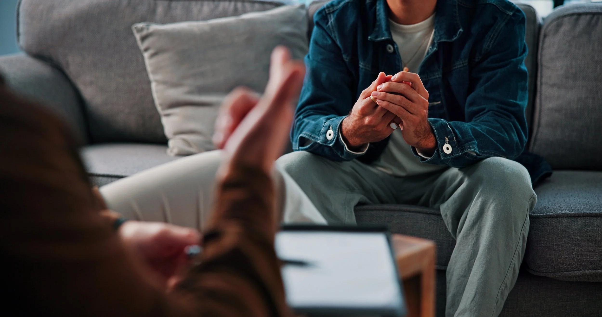 Person sitting on a gray sofa with hands clasped together, wearing a denim jacket and light-colored pants, engaged in conversation, in a cozy indoor setting.