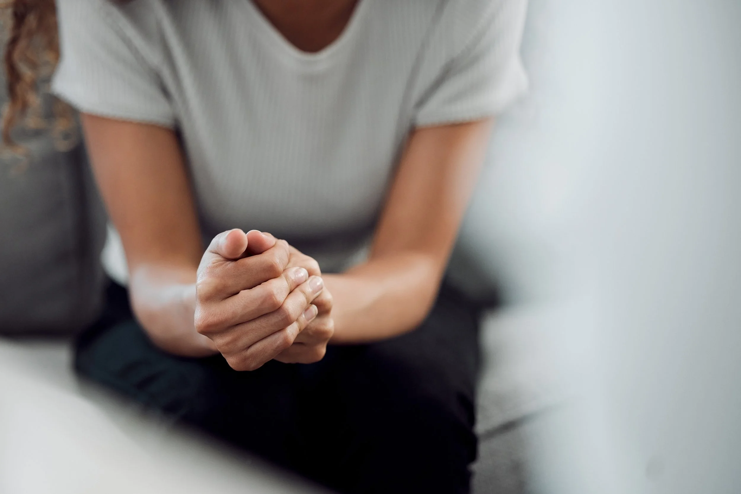 A person wearing a white, ribbed short-sleeved shirt, hands clasped in front, sitting in a relaxed position, sitting in Therapy, with a light-coloured wall in the background.