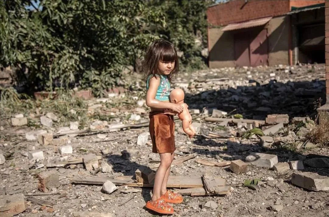 A young girl with long brown hair, wearing a turquoise sleeveless top and brown shorts, is standing outdoors on a rocky, debris-strewn surface. She is holding a small plastic doll close to her chest and looking down with a somber expression.