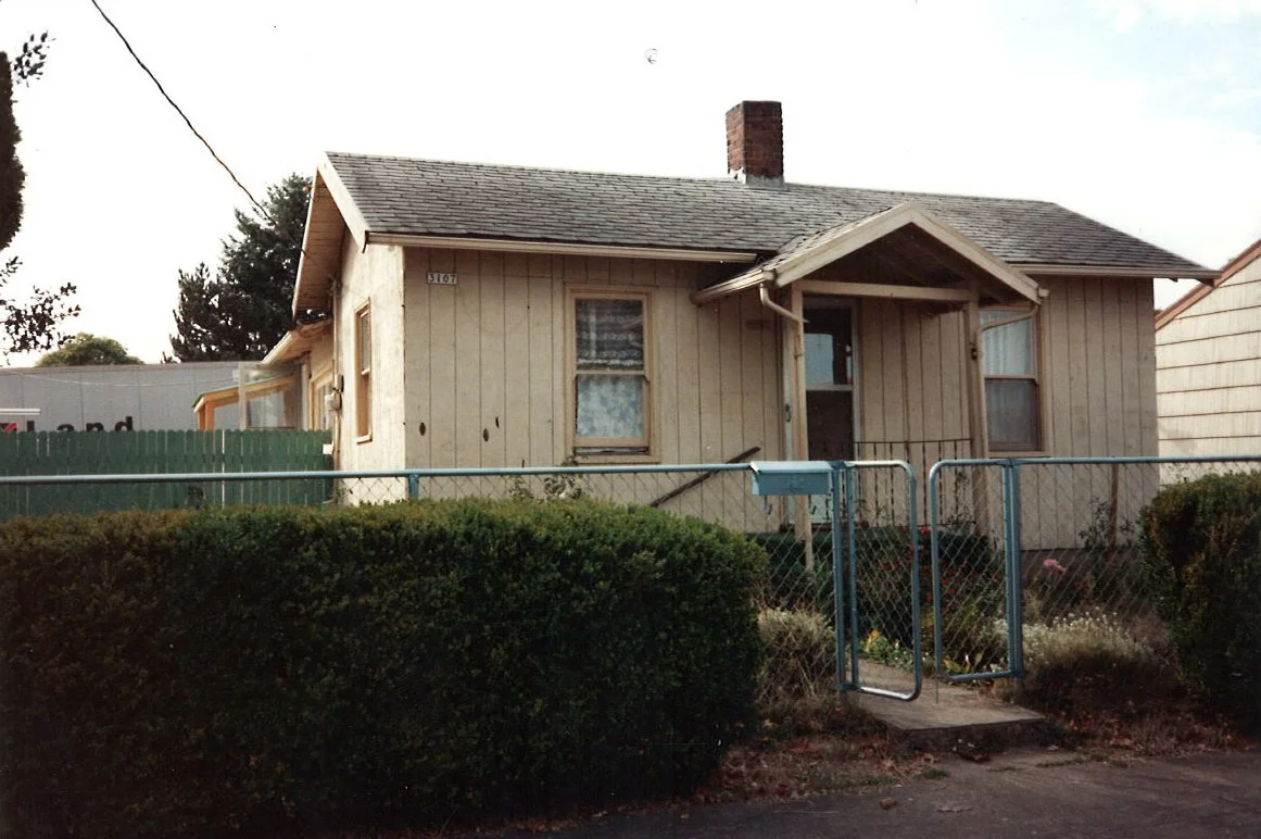 A small, older house with beige vertical siding, a chimney, two windows with curtains, and a front porch with a small overhang, surrounded by a green fence and bushes.