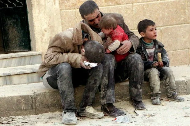 A man sitting on stairs with three children, one of whom is a small boy sitting in the man's lap. The other two children are sitting nearby, all appear dirty and in worn clothing.