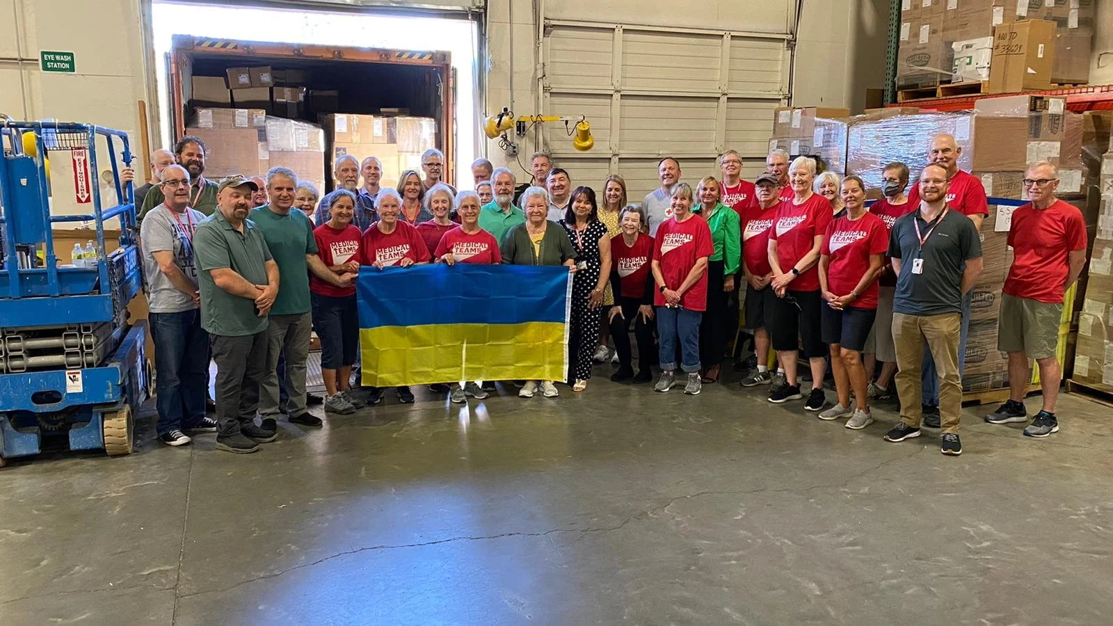 A large group of people, including volunteers in red shirts with 'Medical Teams' written on them, gathered inside a warehouse. Some hold a Ukrainian flag. The warehouse has pallets stored along the walls and a loading dock in the background.