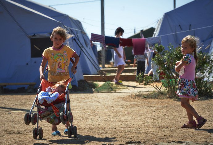 Children playing outside in a refugee camp with tents, some children are walking up steps, one girl is standing with arms crossed, a girl is pushing a stroller with a young child inside.