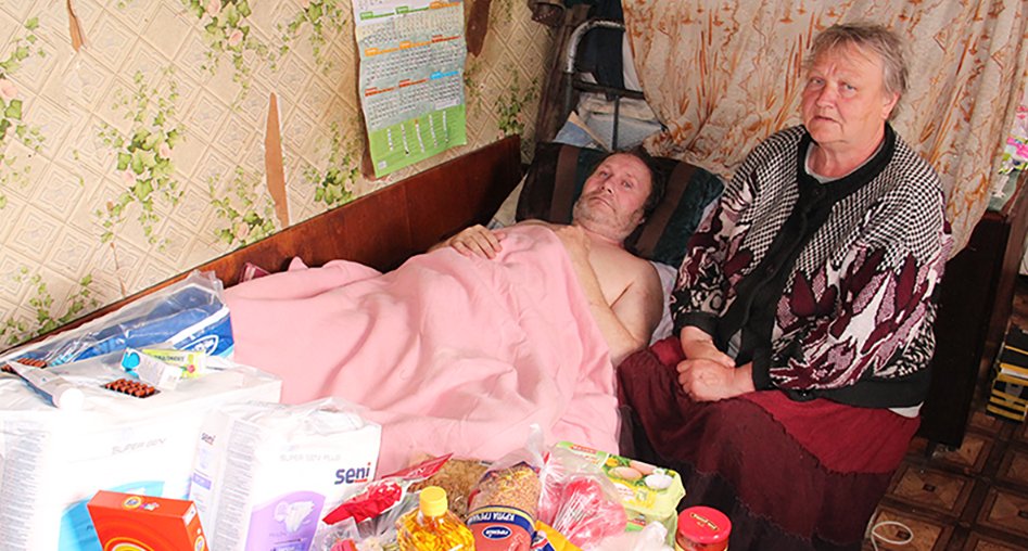 An elderly woman sits beside a man lying in bed, covered with a pink blanket in a cluttered room with patterned wallpaper and various items on a table.
