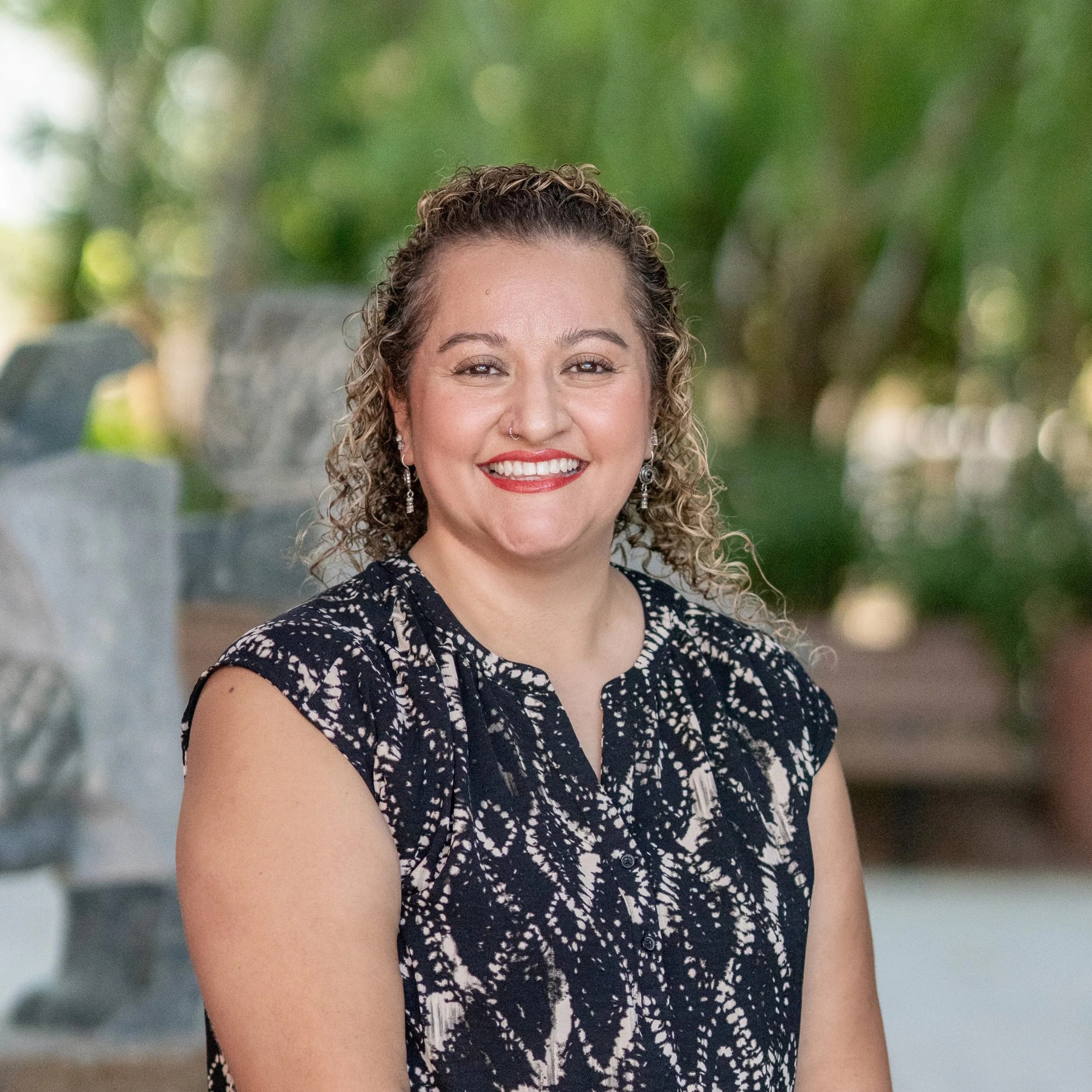 Hispanic woman standing outside with long curly hair, dangling earrings, and red lipstick. Wearing a black dress shirt with tan lines.