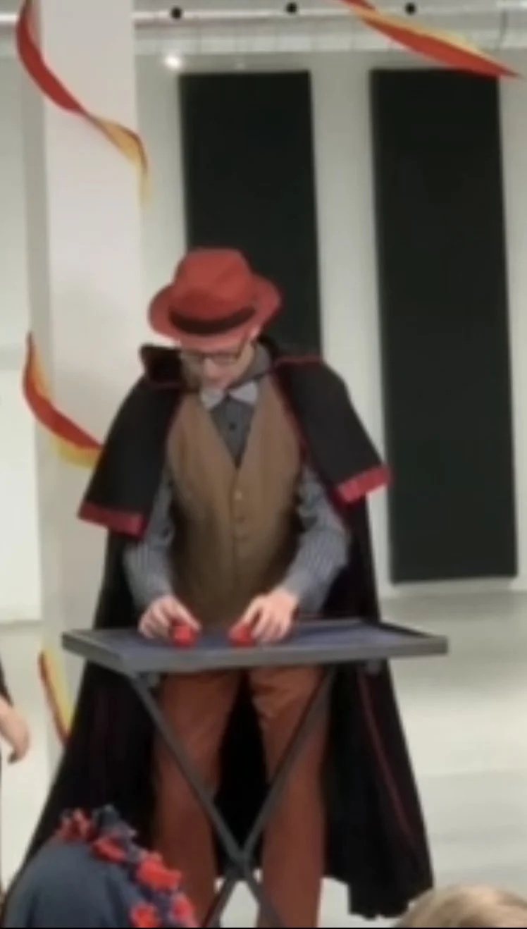Justin, dressed in a cape, brown vest, and fedora hat, performing magic with two red balls at a table in a festive setting with red and orange decorations.