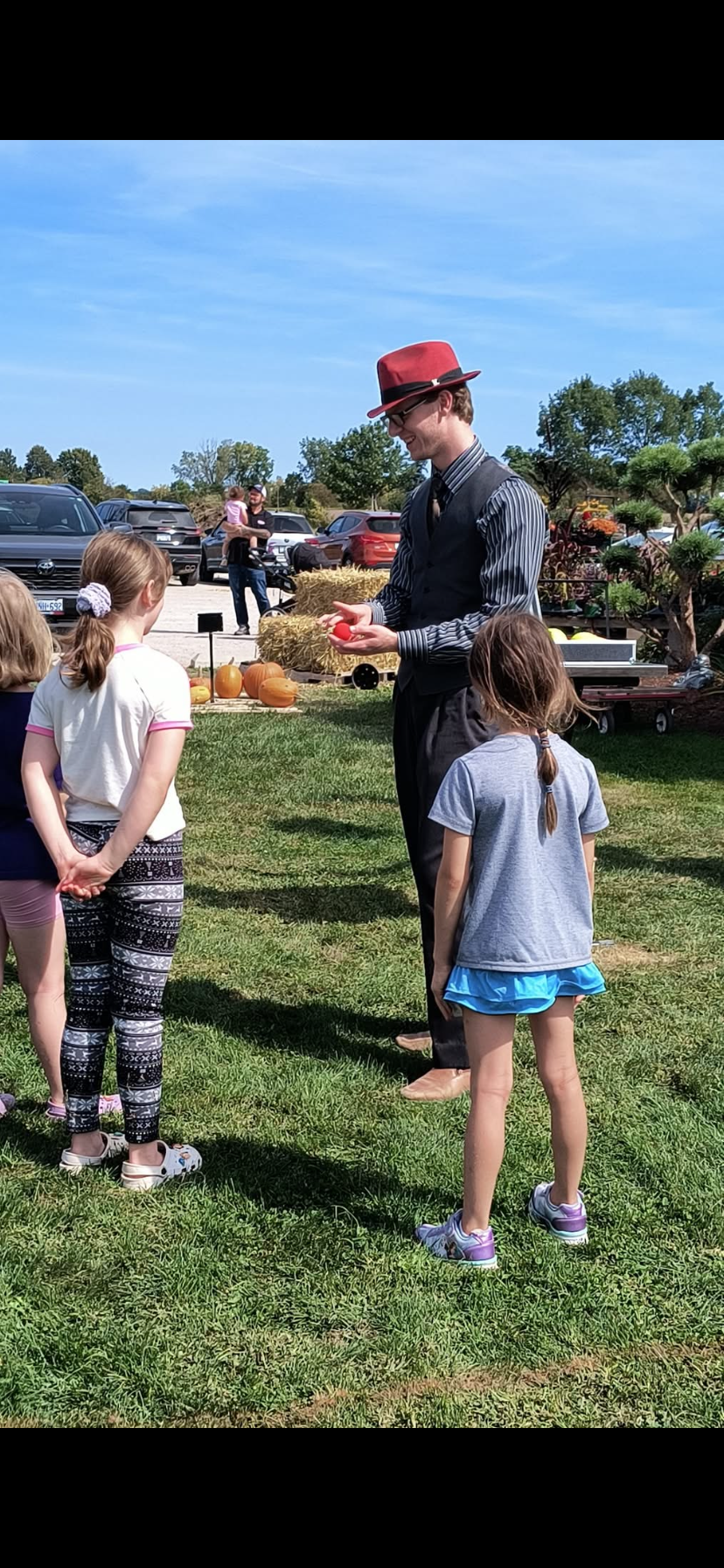Justin, in a red fedora, glasses, and a striped shirt performs a magic trick for a group of children in an outdoor fair area, with pumpkins and decorated trees in the background.