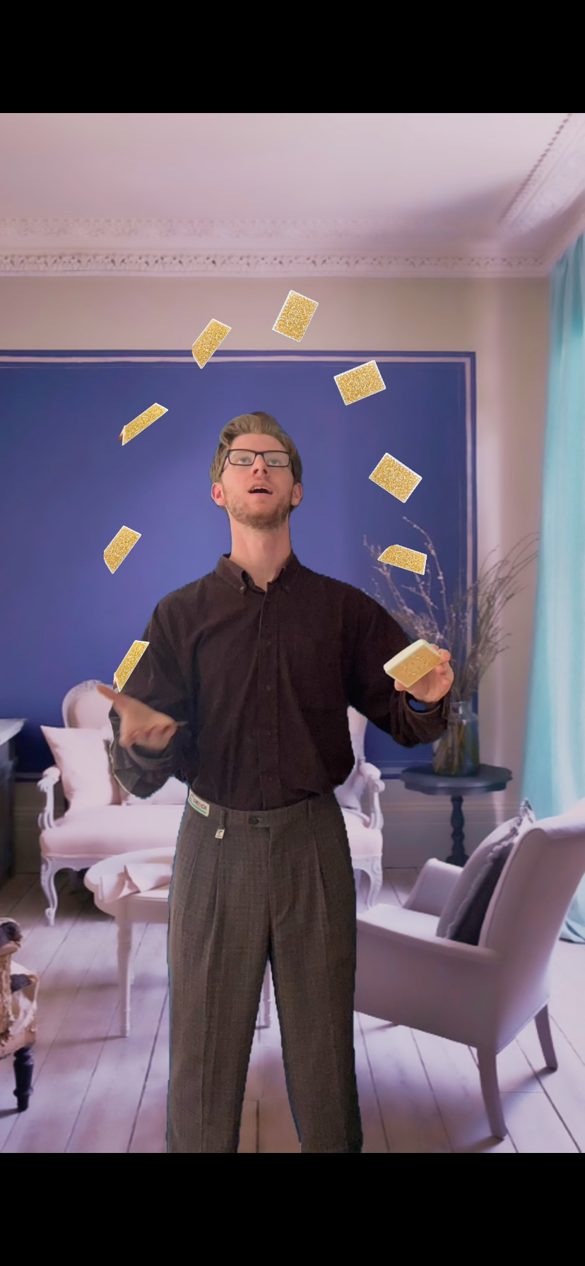 Justin, wearing a black shirt and patterned pants, juggles virtual glittering cards in a furnished living room.