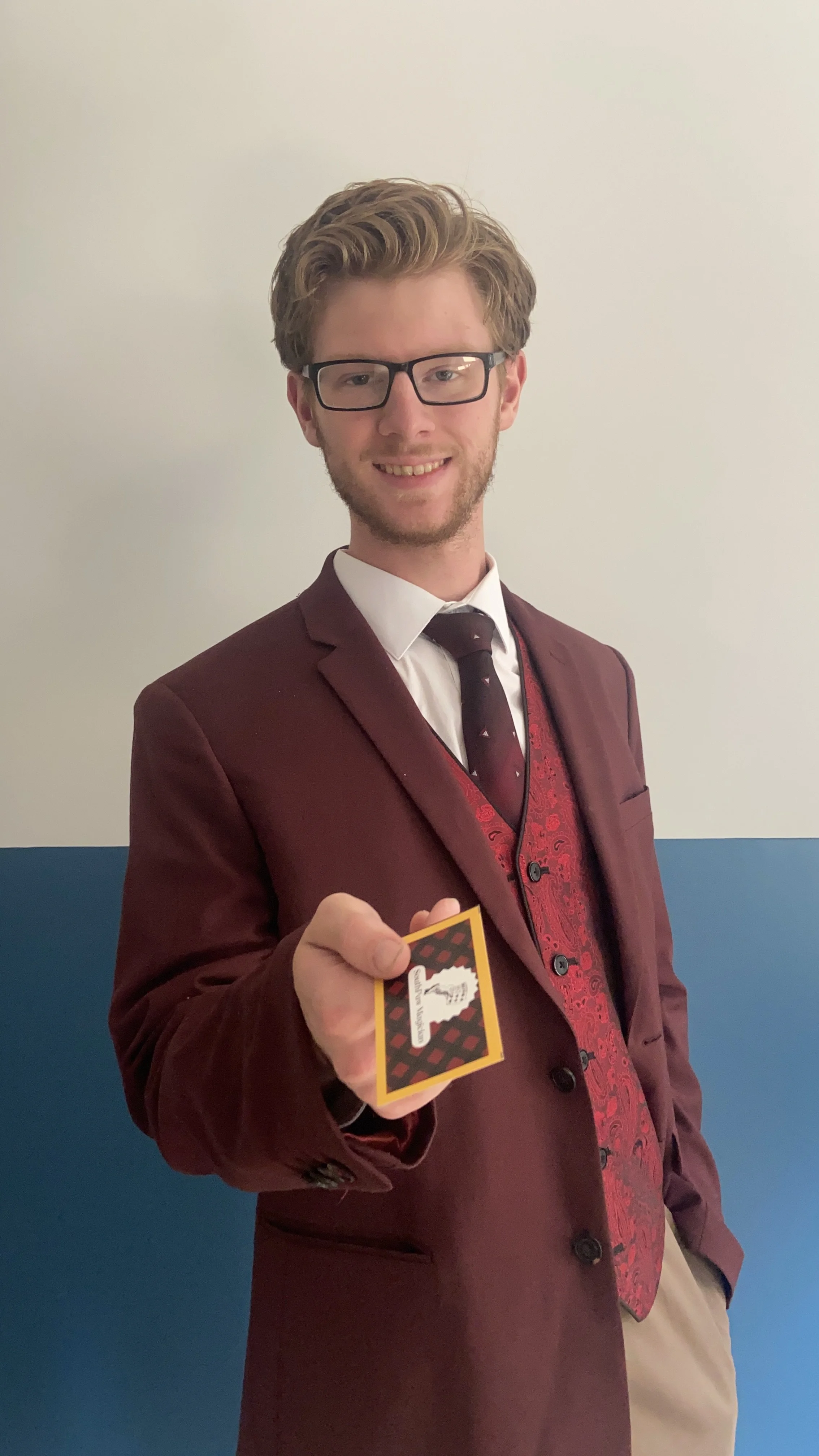 A young man with glasses, wearing a maroon suit, red vest, white shirt, and black tie, is smiling and holding out a card towards the camera.