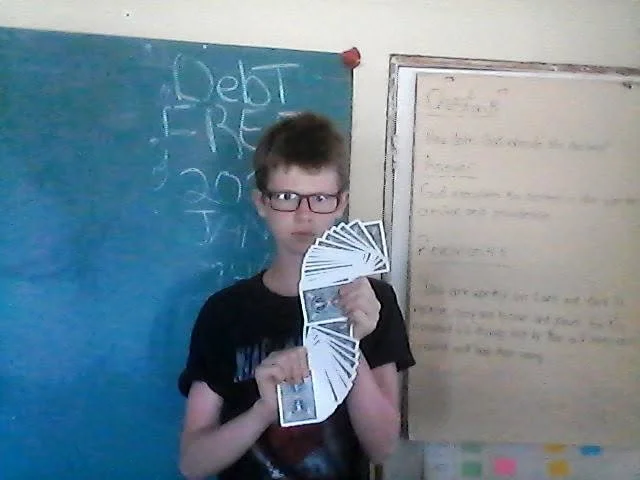 A young boy with glasses and a black T-shirt holding a spread-out fan of playing cards, standing in a classroom with a blue chalkboard and a whiteboard behind him.