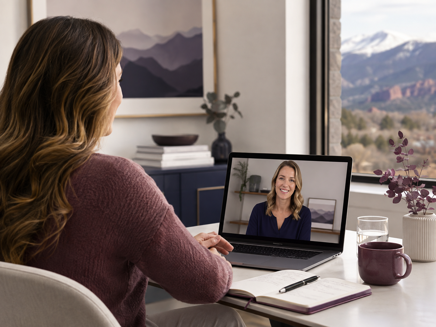 Woman with long wavy hair on a video call, sitting at a desk with a notebook, pen, mug, glass of water, and a potted plant, with a window showing mountains in the background.
