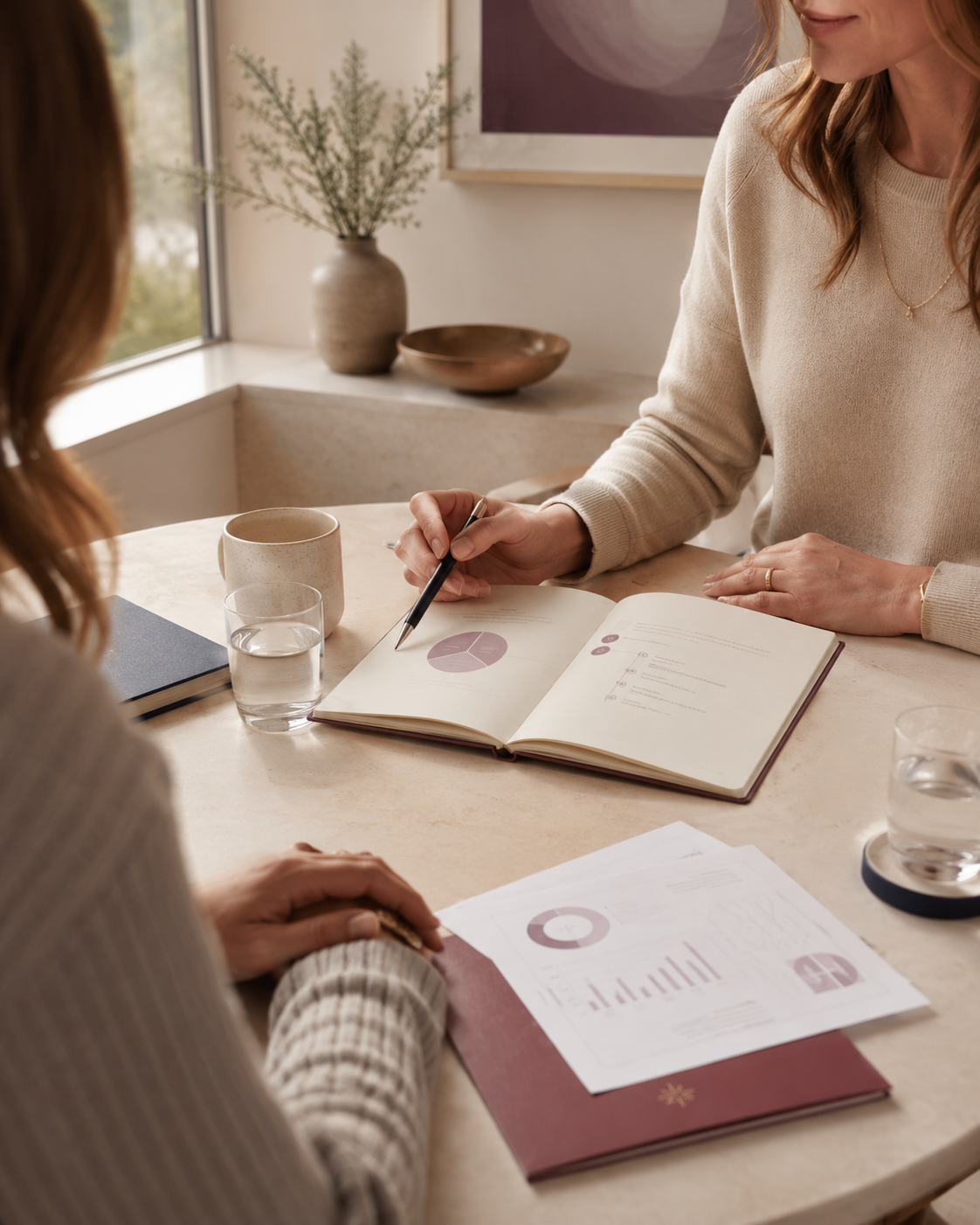 Two women having a meeting at a table with documents, notebooks, and drinks in a bright room with a window and decorative vases in the background.