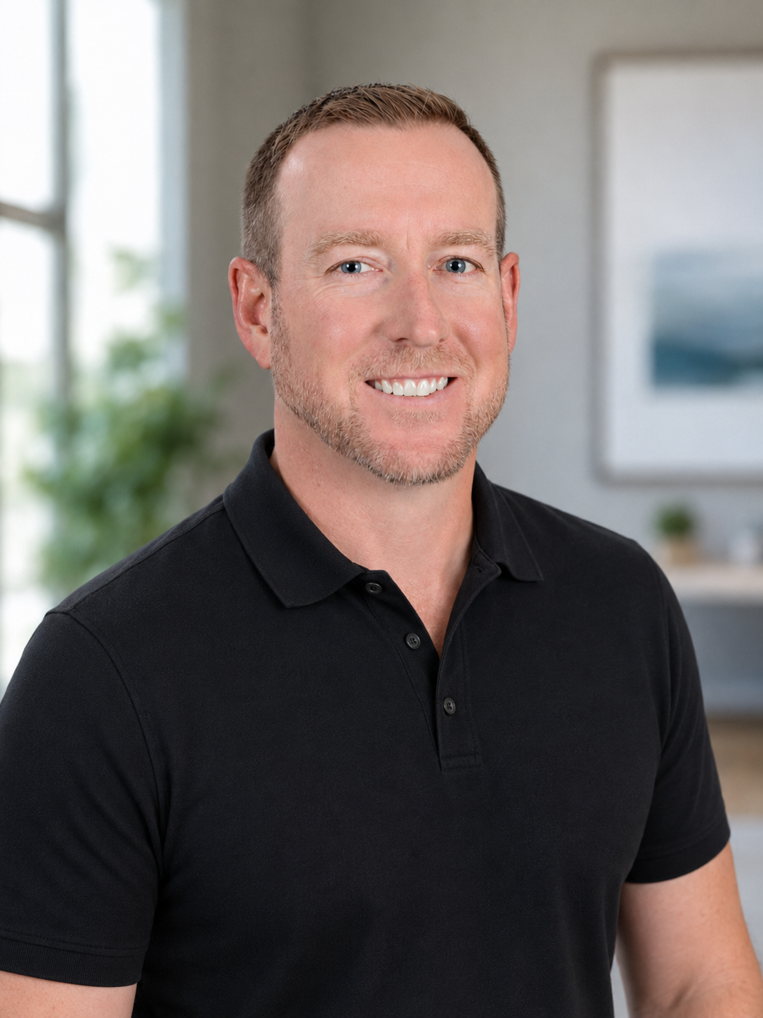 A man with short, light brown hair, a beard, and blue eyes, smiling, wearing a black polo shirt, indoors with a window, plant, and framed artwork in the background.