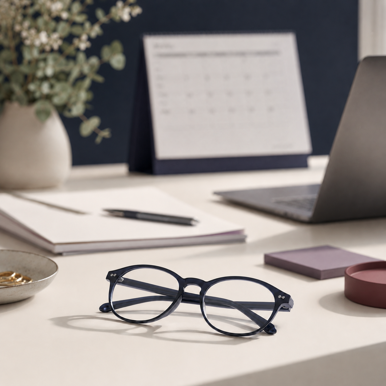 Eyeglasses on a desk with a notebook, pen, and plant.