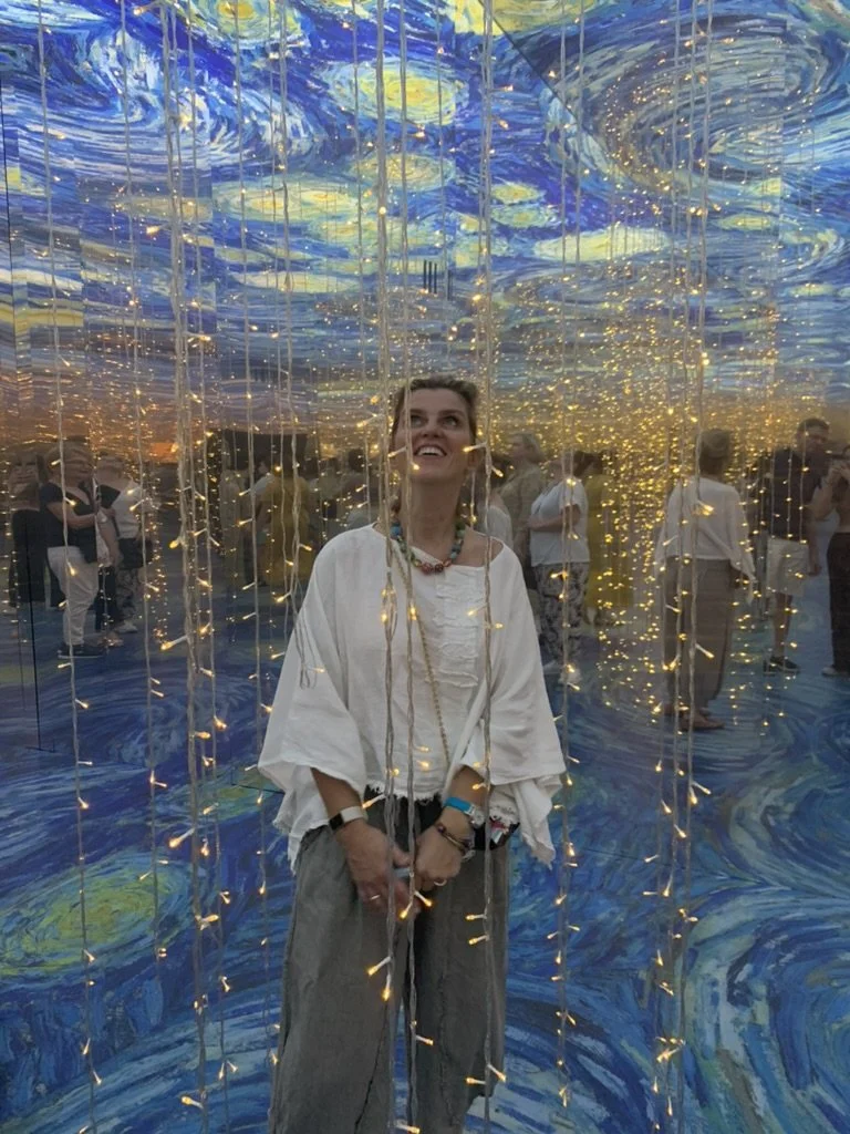 A woman smiling inside a reflective art installation with swirling blue patterns and hanging string lights, with several people in the background at an art exhibit.