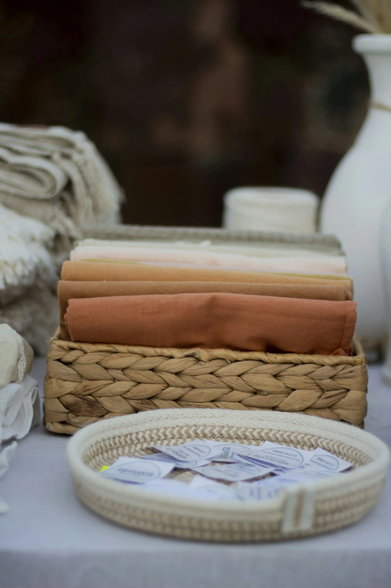 A display of folded fabric napkins in a woven basket with tags, and nearby croissant-patterned linen and white dishes in the background.