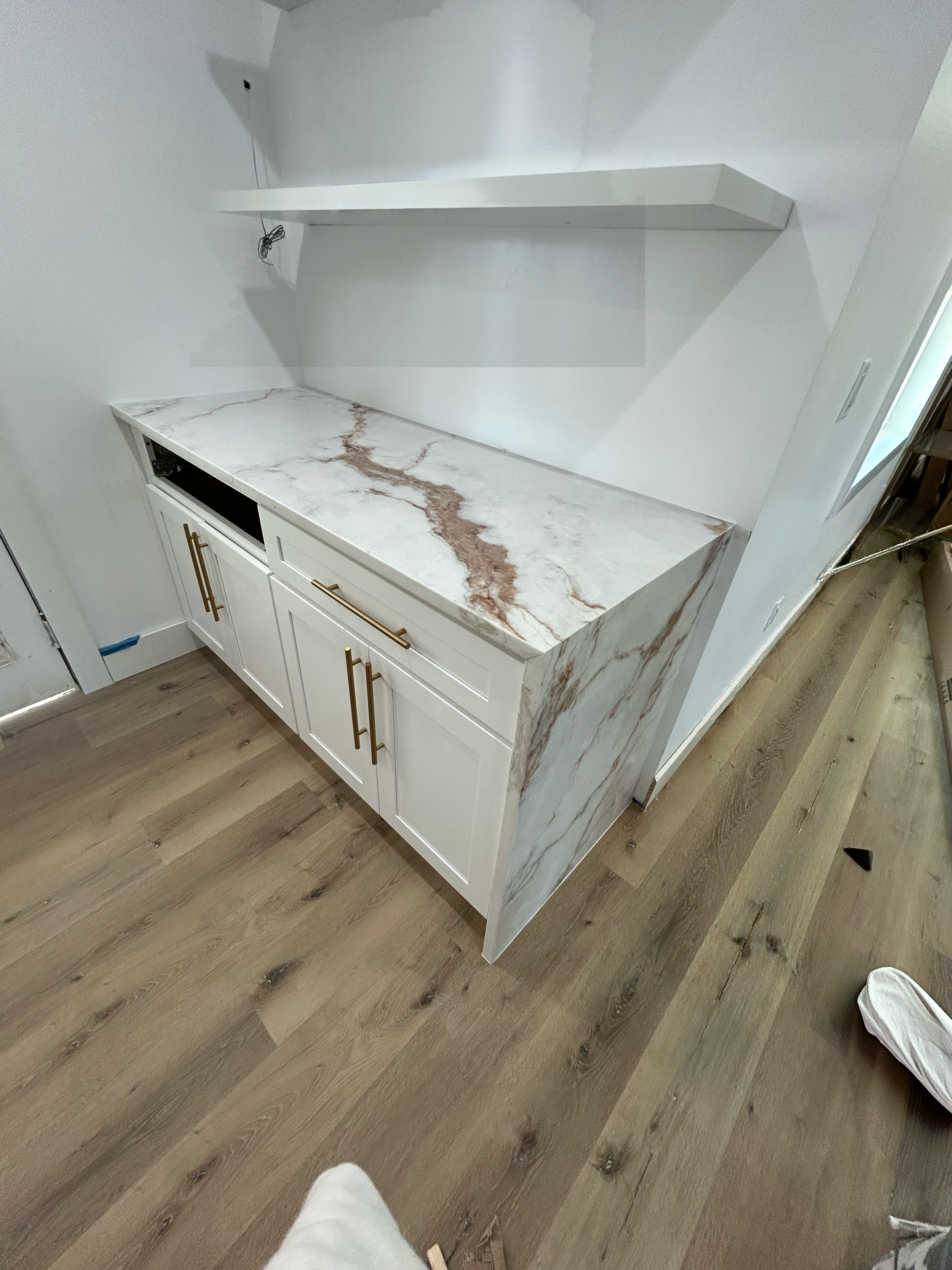 Interior view of a kitchen with white cabinetry, marble countertop with brown and beige veins, wooden floor, and a white wall with an empty shelf.