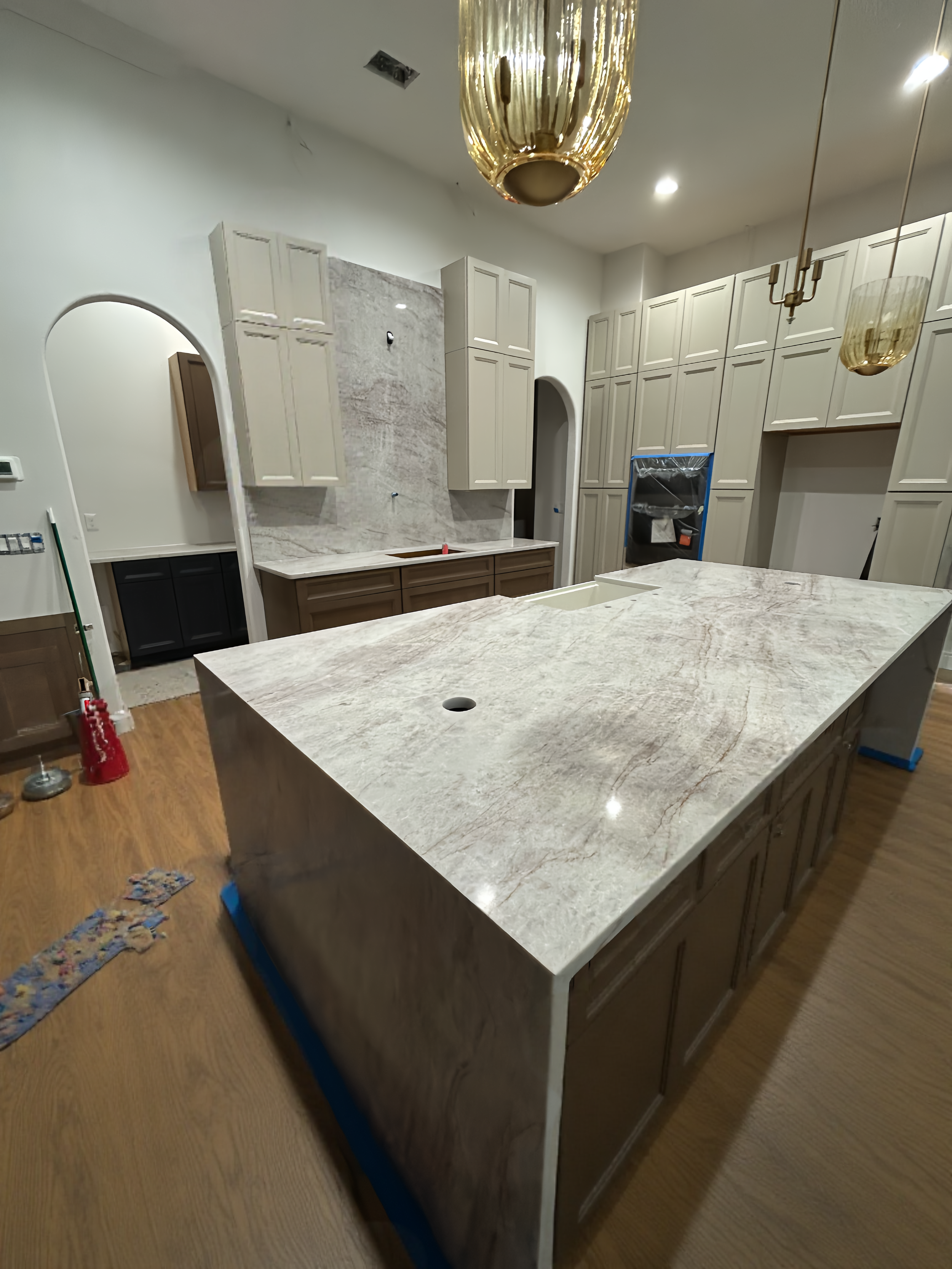 A kitchen under renovation with a large marble island in the foreground, cream-colored cabinets, and pendant lights hanging from the ceiling.