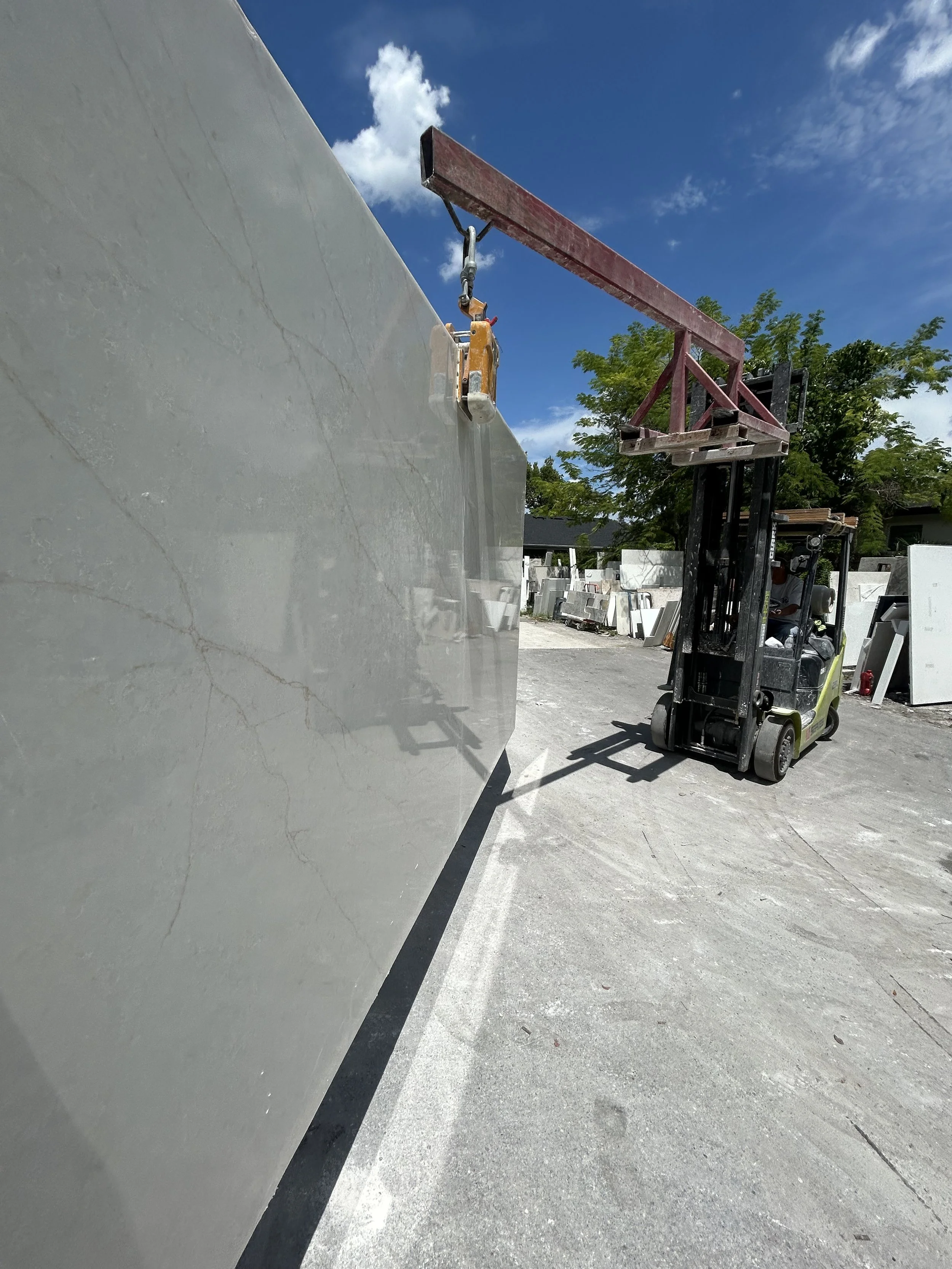 A forklift lifting a large marble slab outdoors under a blue sky with some clouds, with other marble slabs and green trees in the background.