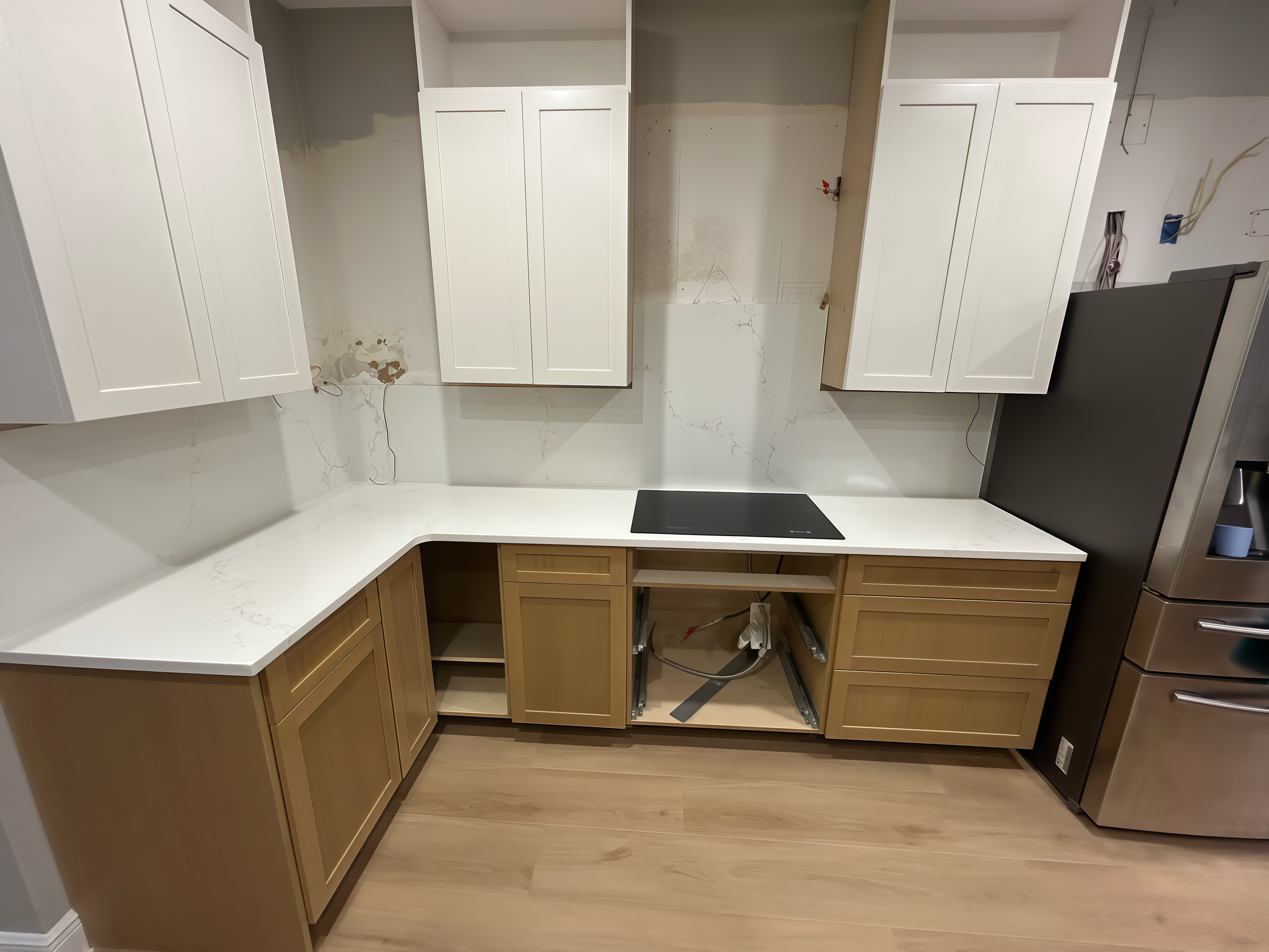 Kitchen cabinets with missing doors, countertop, and a stovetop installed. Walls have some damage and wiring open. The lower cabinets are light wood, and there is a tiled floor.