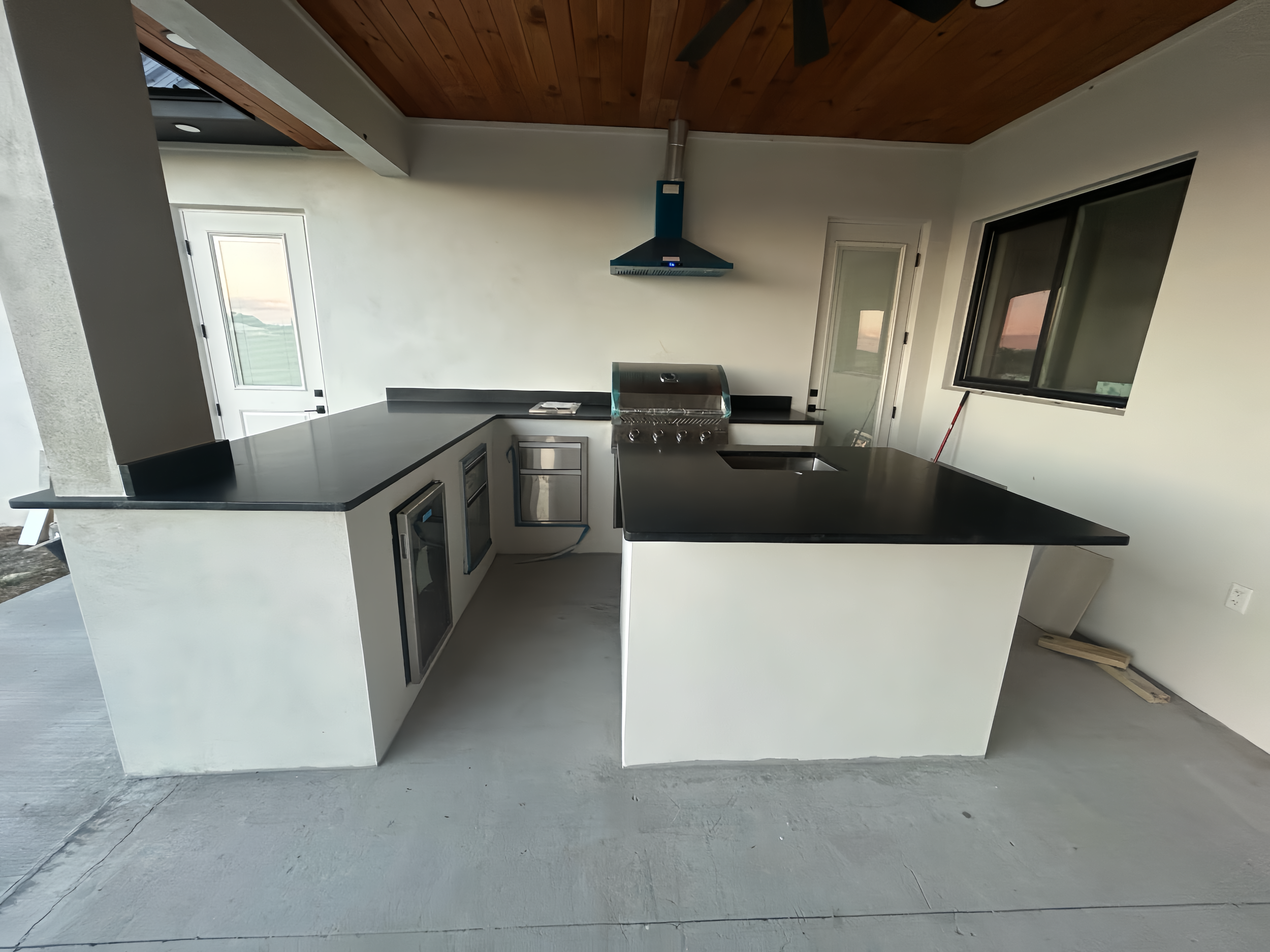 Modern kitchen with black countertops, a stainless steel grill, and a black range hood. White walls, a window, a door, and a wooden ceiling are visible.