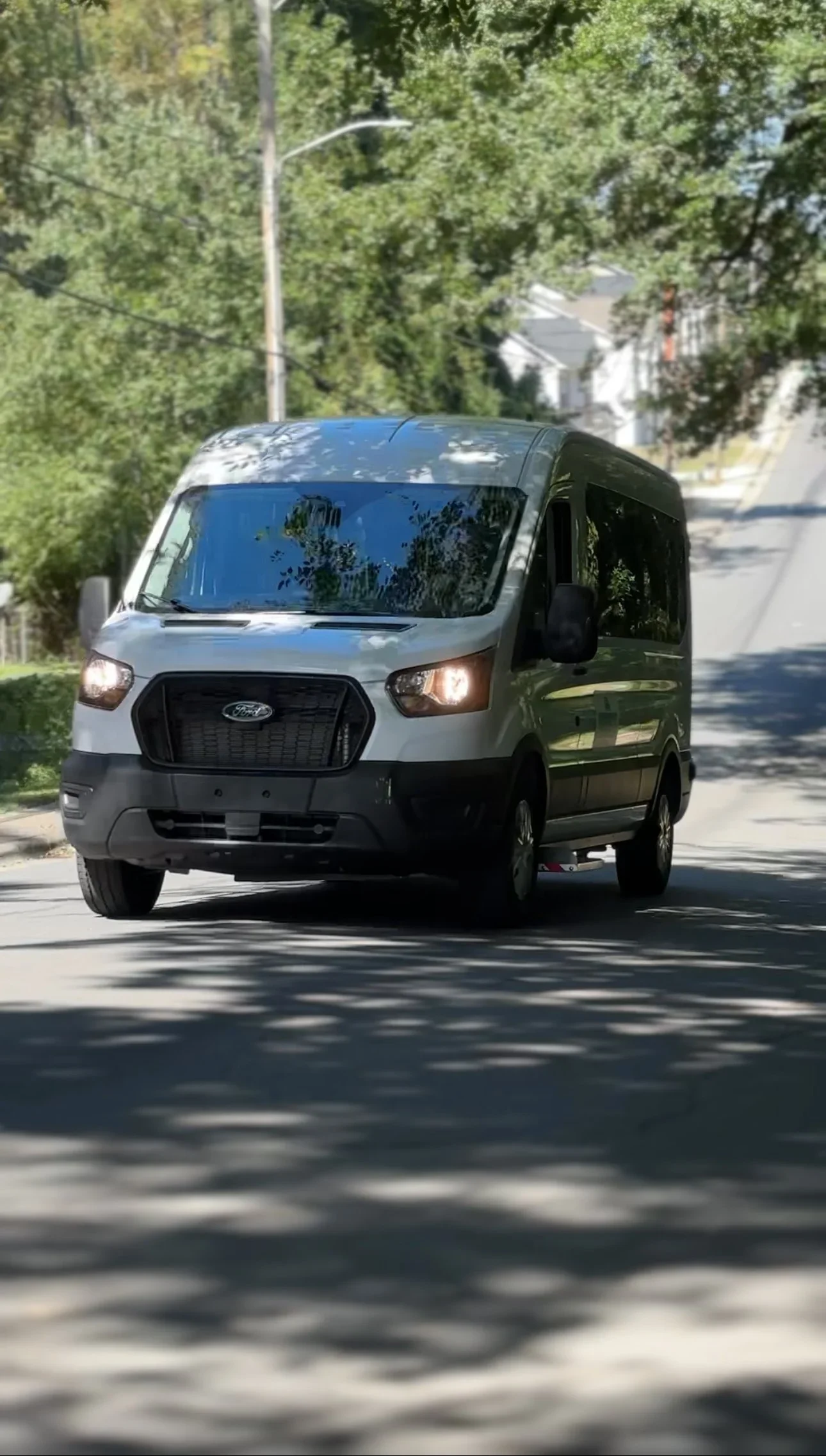 A white Ford cargo van driving on a tree-lined residential street during daytime.