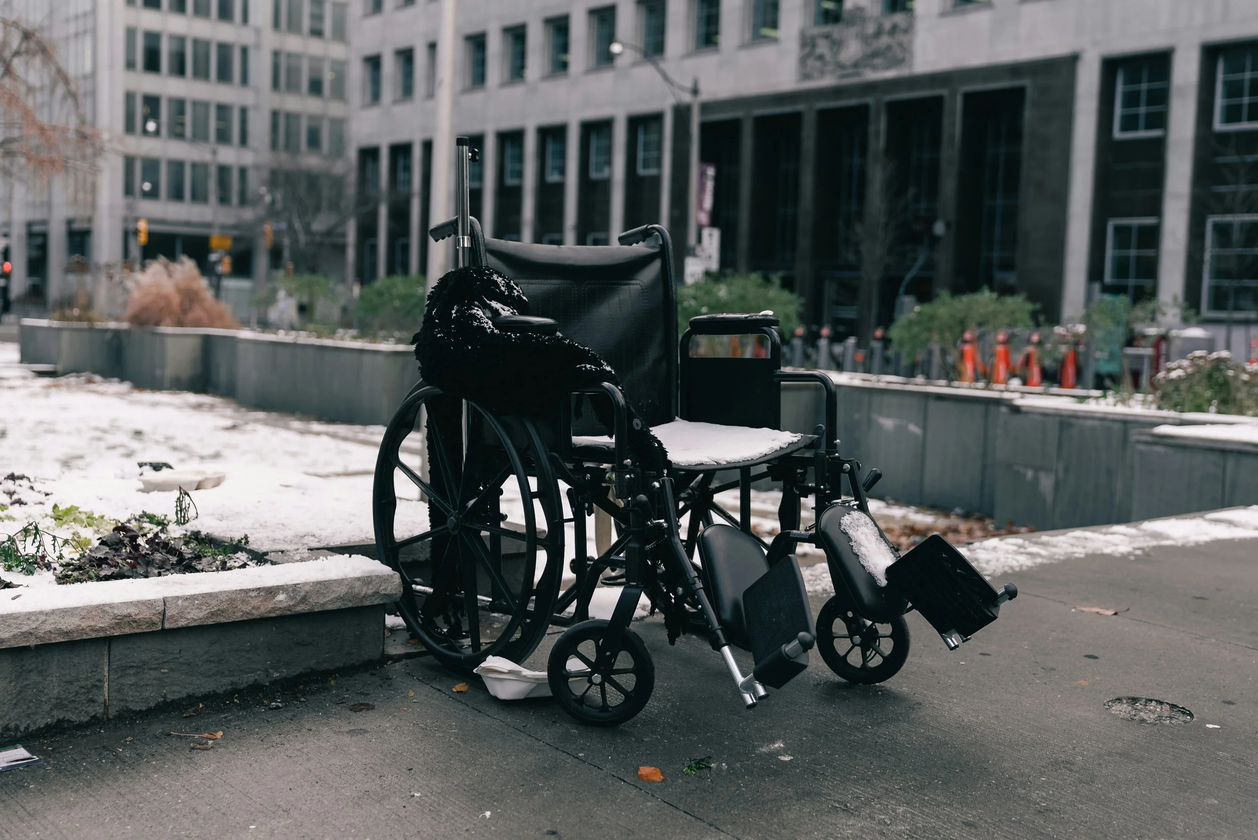 An empty wheelchair parked on the sidewalk in an urban area with snow on the ground, surrounded by buildings and some plants.