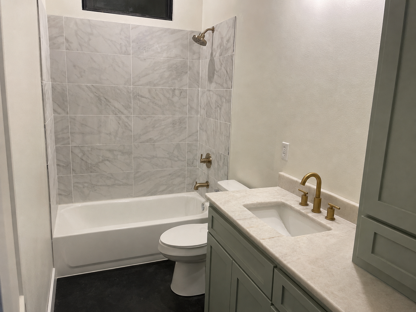 A modern bathroom featuring a bathtub with a marble-tiled wall, a toilet, and a vanity with a marble countertop and gold fixtures.