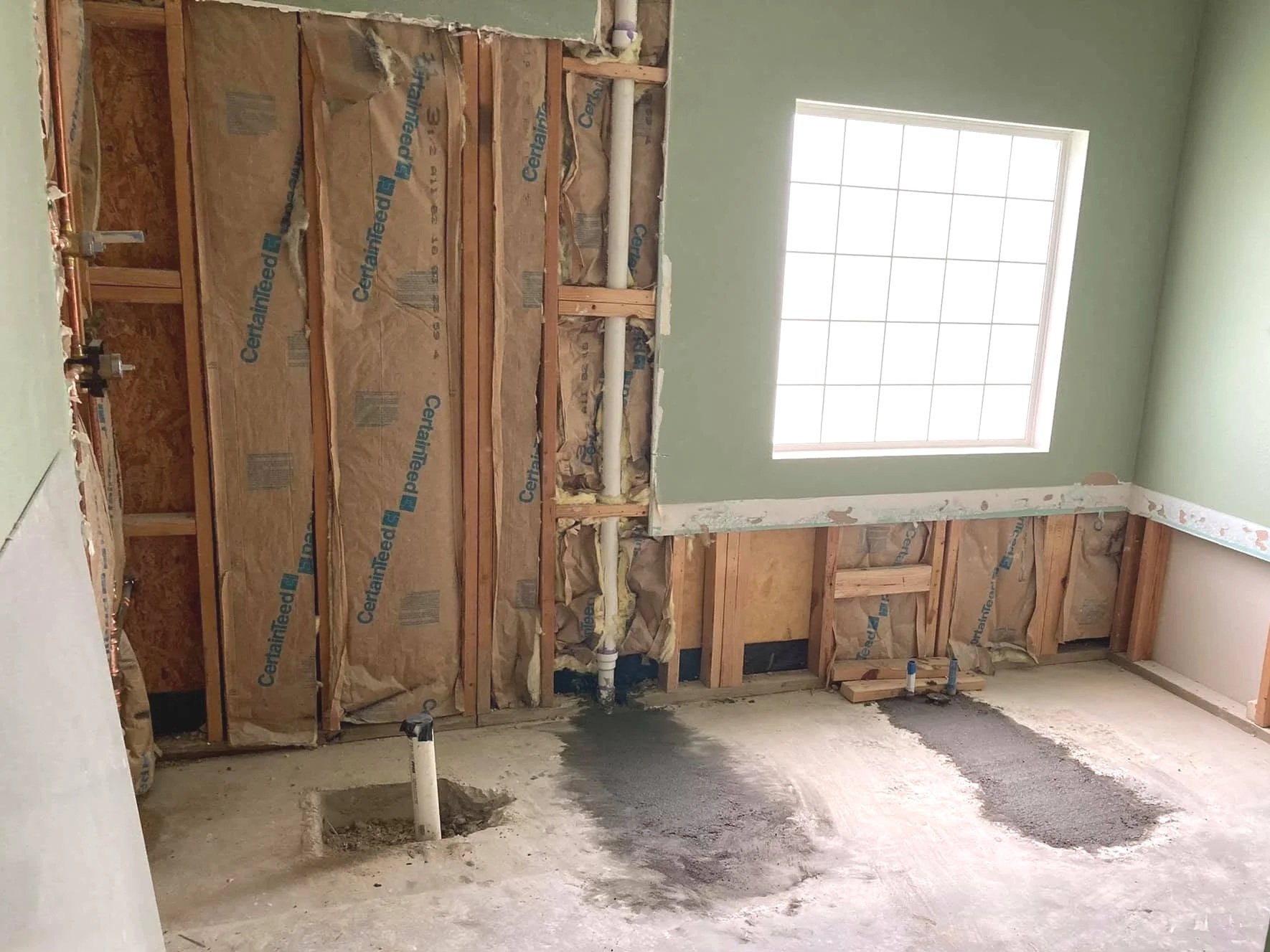 Interior of a room under renovation with exposed wall studs, plumbing pipes, and insulation. There is a window on the right wall and a floor with construction dust and holes for future fixtures.