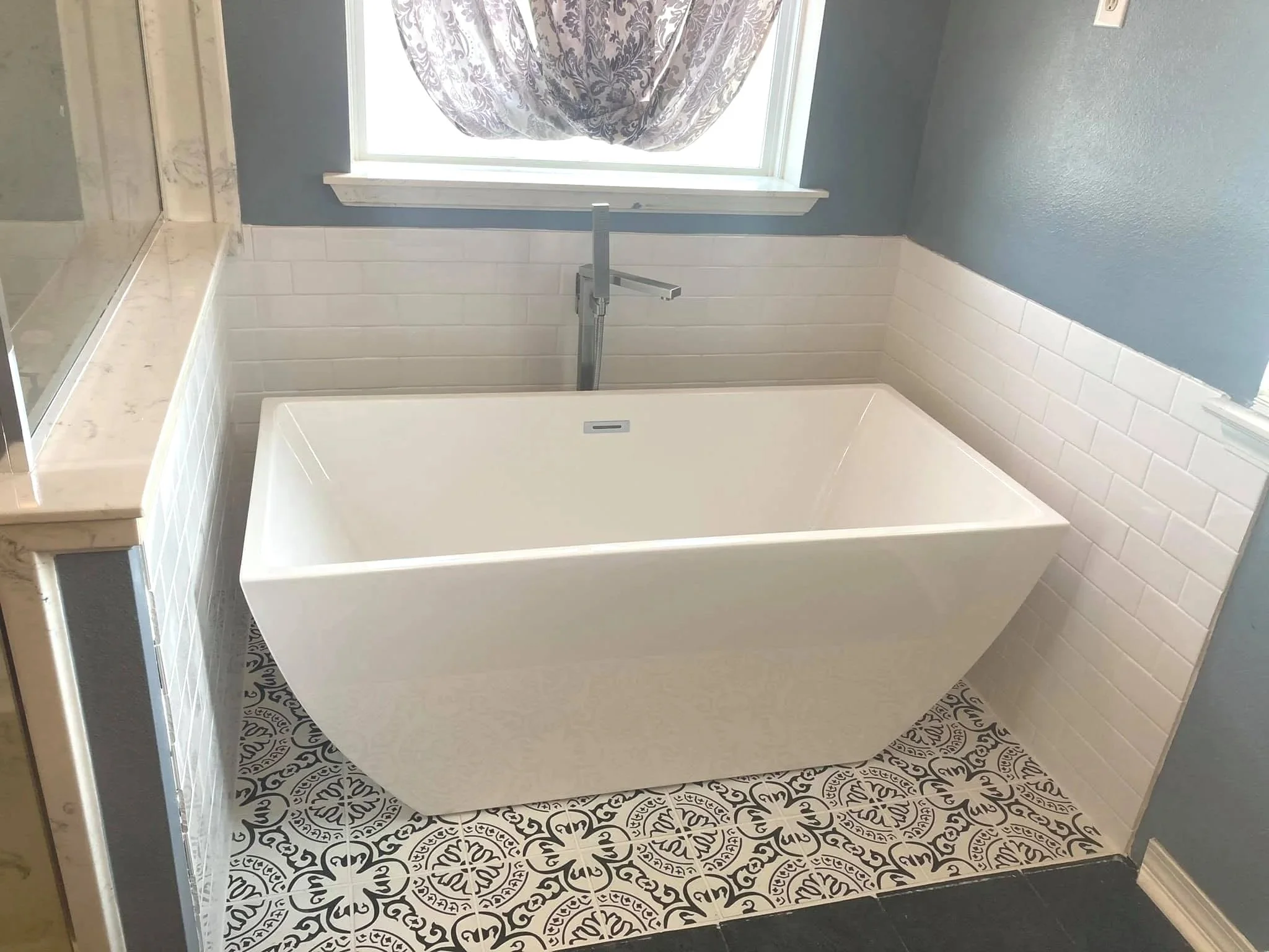 Bathroom with patterned floor tiles, a white bathtub, a blue wall, a window with a curtain, a marble countertop, and white tiled walls.
