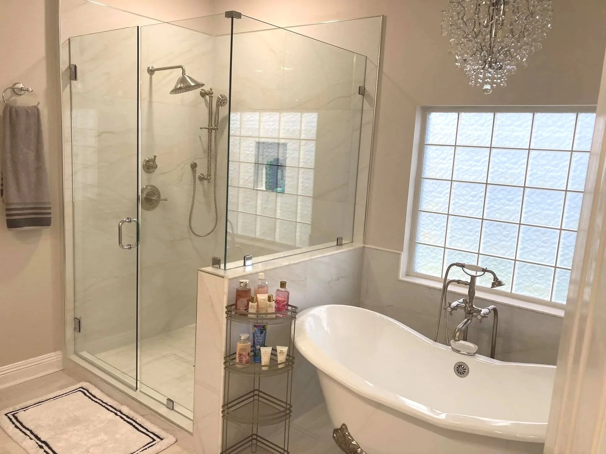 Bathroom with a glass shower enclosure, marble walls, a large bathtub, a chrome chandelier, and a window with frosted glass.