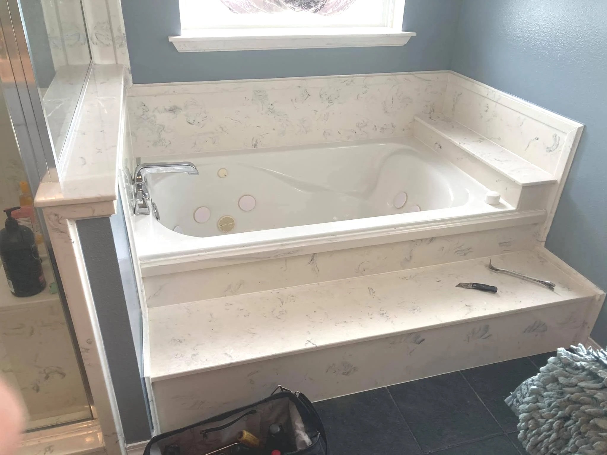 A bathroom with a whirlpool bathtub surrounded by beige marble-like panels. A window above the bathtub provides natural light. Tools and supplies laid out around the bathtub suggest ongoing installation or renovation work.