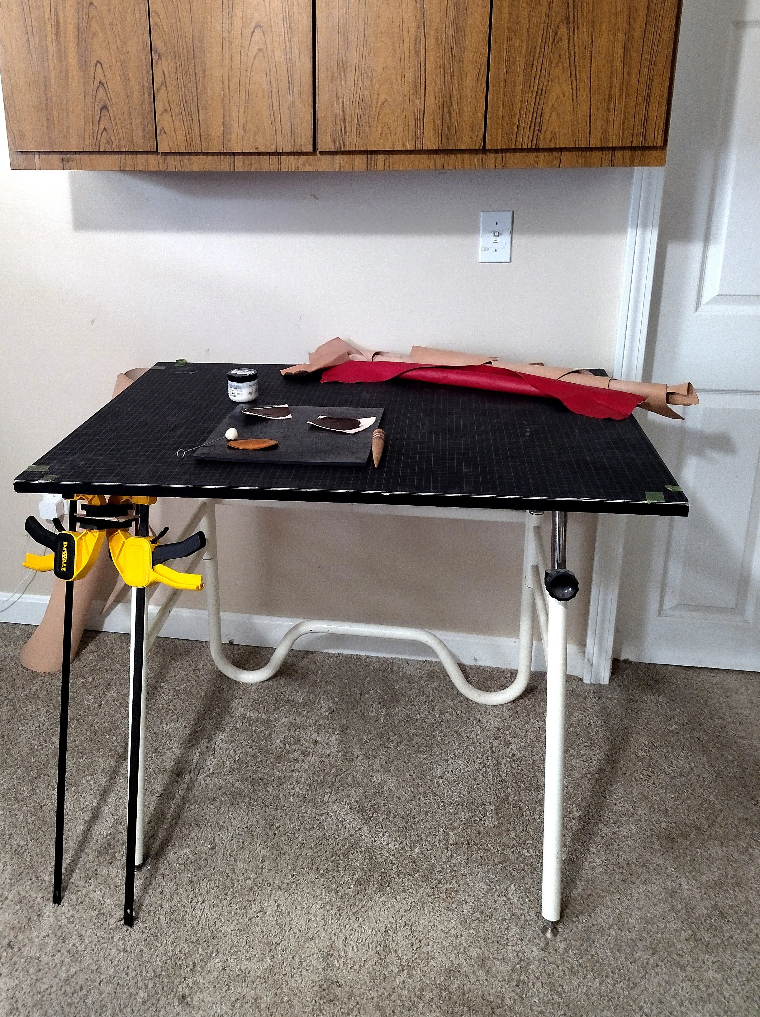A picture of a drafting table covered in a cutting mat. Rolls of leather lay atop it and rest against it. On the table also sits various tools used in leatherworking along with leather pieces that have been recently stained.