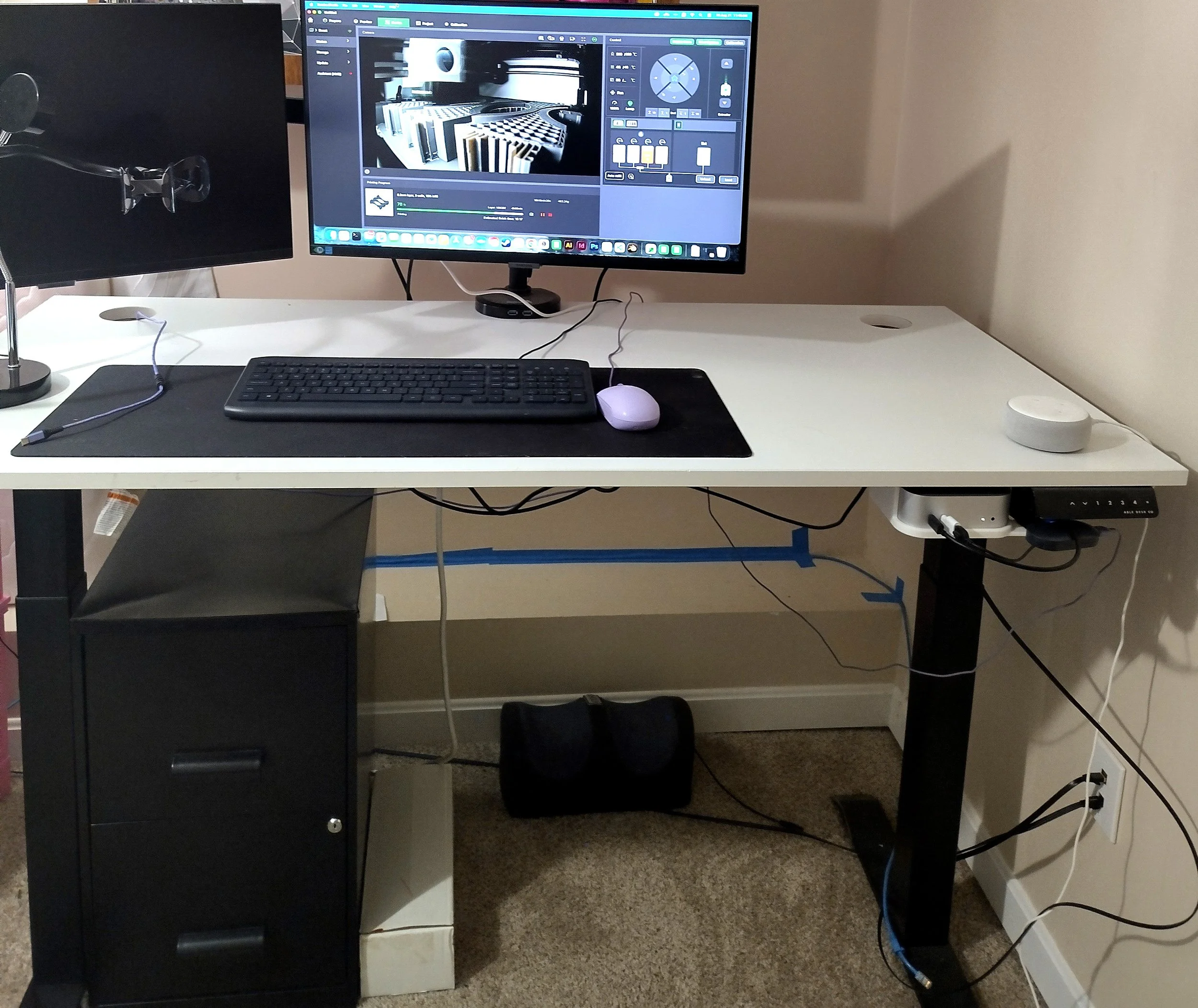 A picture of a standing desk with two monitors. One monitor is on, displaying the live video feed of a 3D printer running.