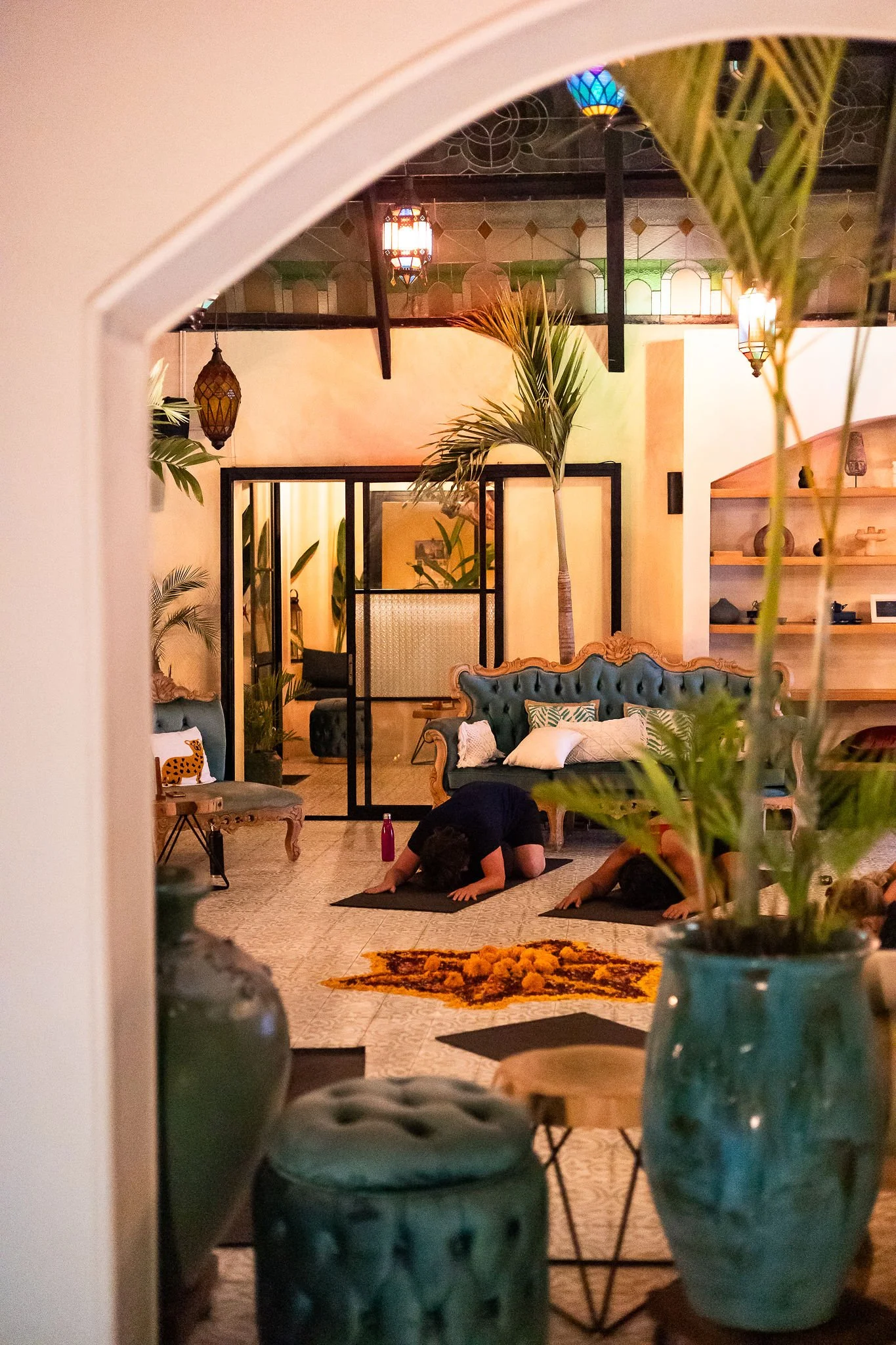 People practicing yoga in a cozy, well-lit room decorated with plants, colorful hanging lanterns, and shelves with decorative items.