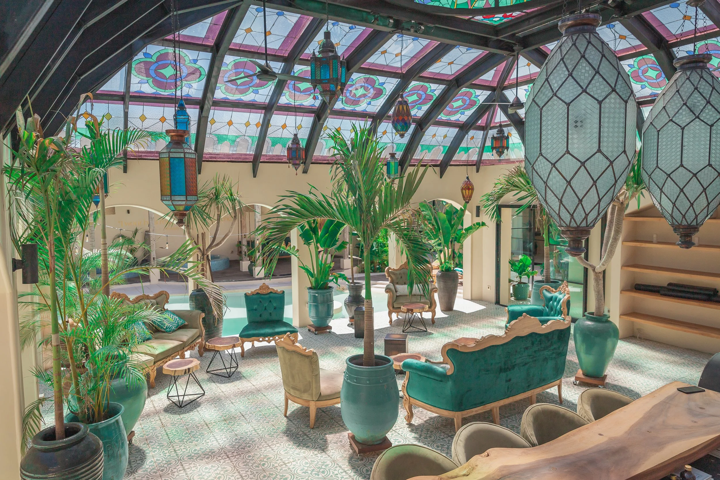 Indoor space decorated with green plants, teal and beige furniture, and hanging Moroccan-style lanterns under a stained-glass ceiling.