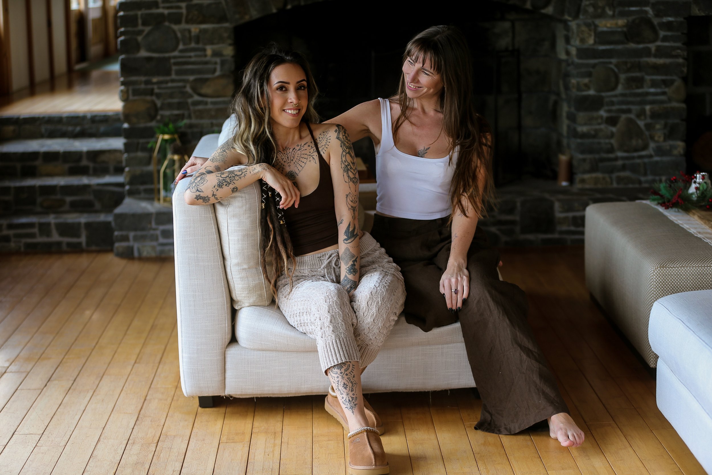 Two women sitting on a beige couch in a living room with a stone fireplace in the background. One woman with long dark hair and tattoos on her arms is smiling at the camera. The other woman with long brown hair is smiling and looking at her friend, with her arm around her. The blonde woman is wearing a white tank top and dark pants, while the other woman is wearing a brown top and beige pants. There are holiday decorations on the table nearby.