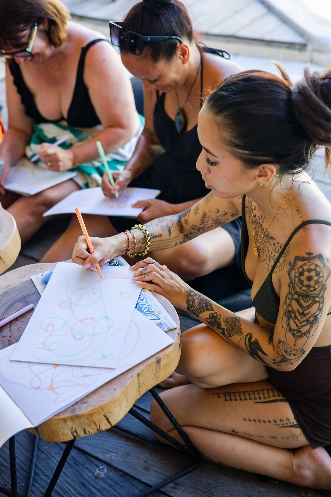 Three women sitting at a wooden table, drawing on papers with colored pencils, in an outdoor setting with a sunlit background.