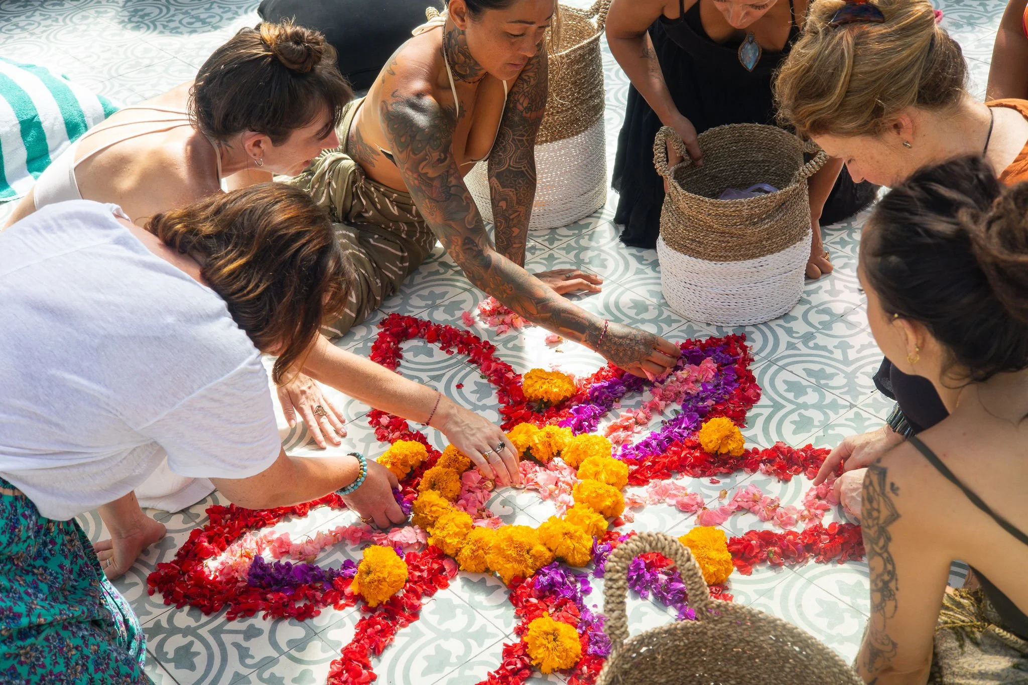 Group of women gathered around laying flower petals on a patterned outdoor floor, forming a heart shape with vibrant orange, pink, purple, and red flowers.