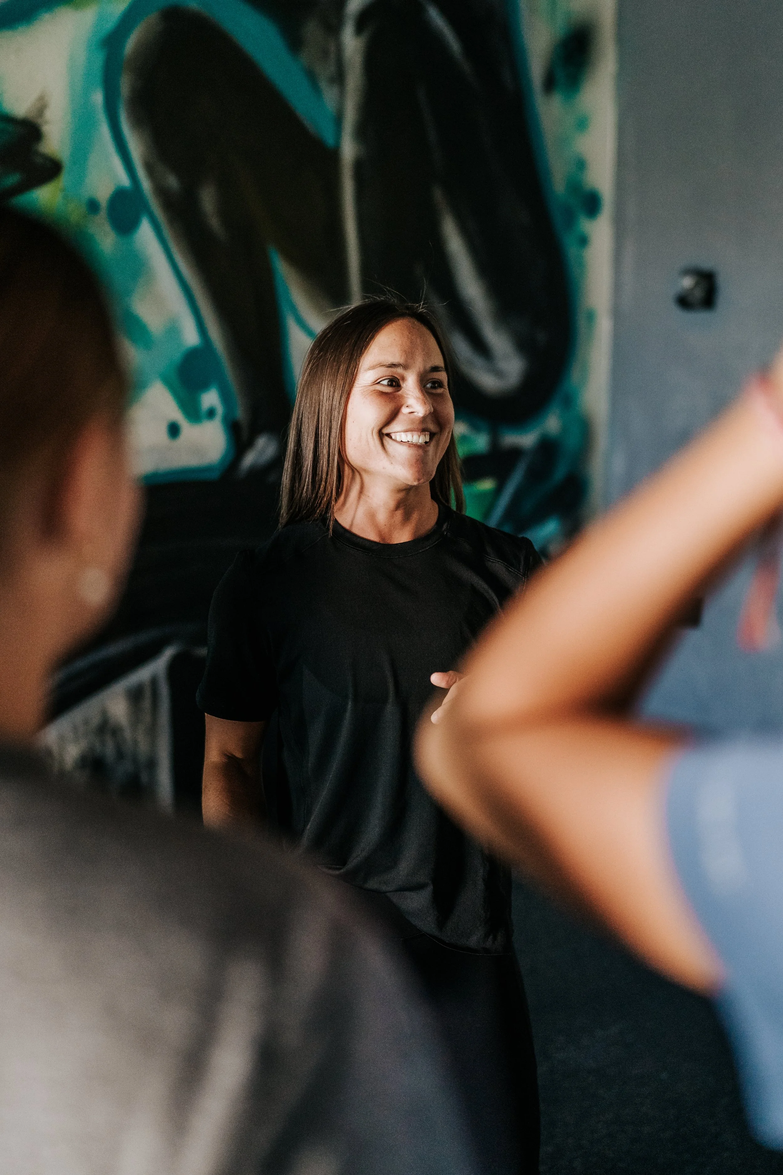 A woman with brown hair smiling in front of a graffiti wall, with blurred hands in the foreground.